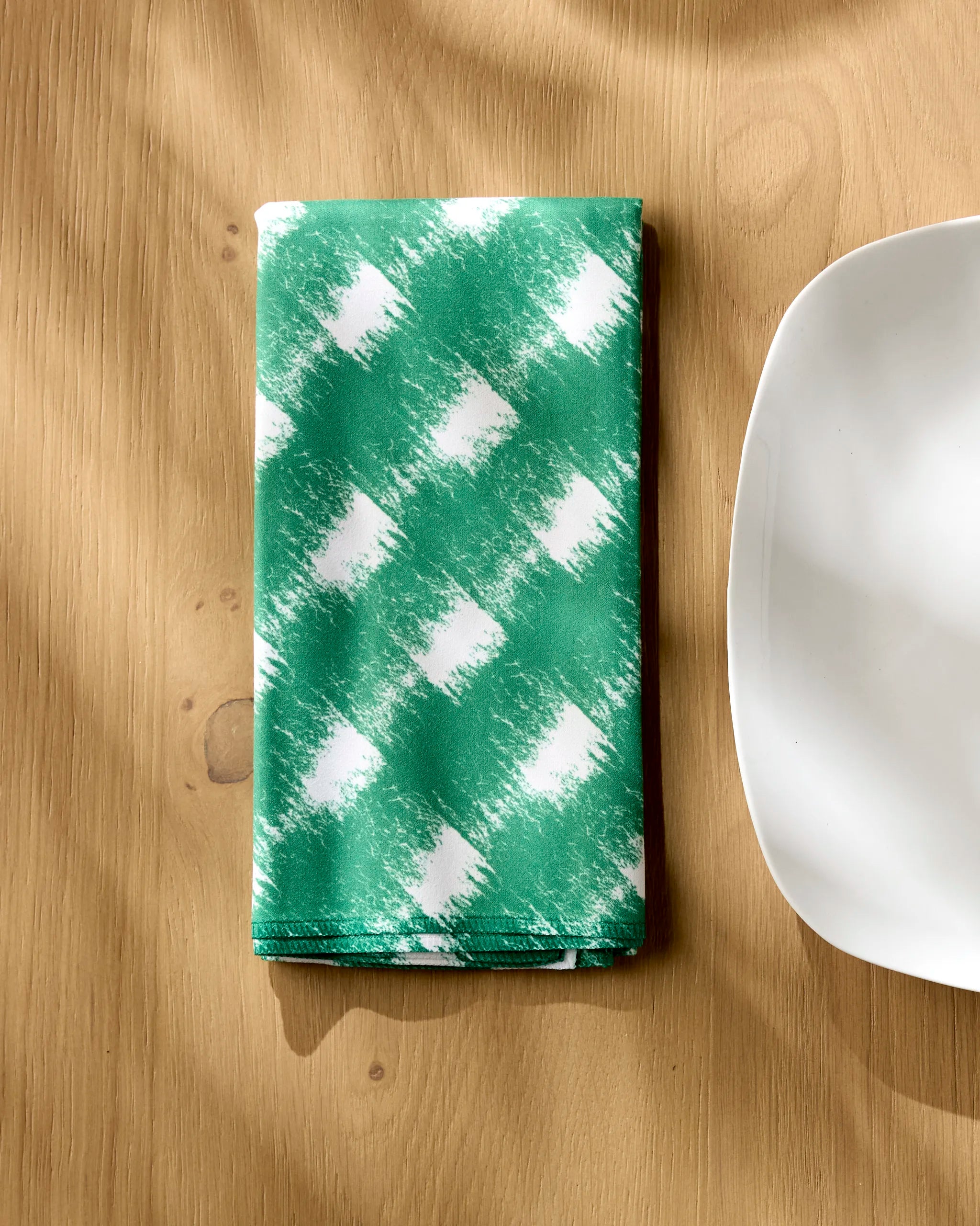 A Geometry Hairy Check Green absorbent cloth napkin with a green and white pattern is neatly folded on a light wooden table beside the edge of a white square plate, soft shadows across the scene.