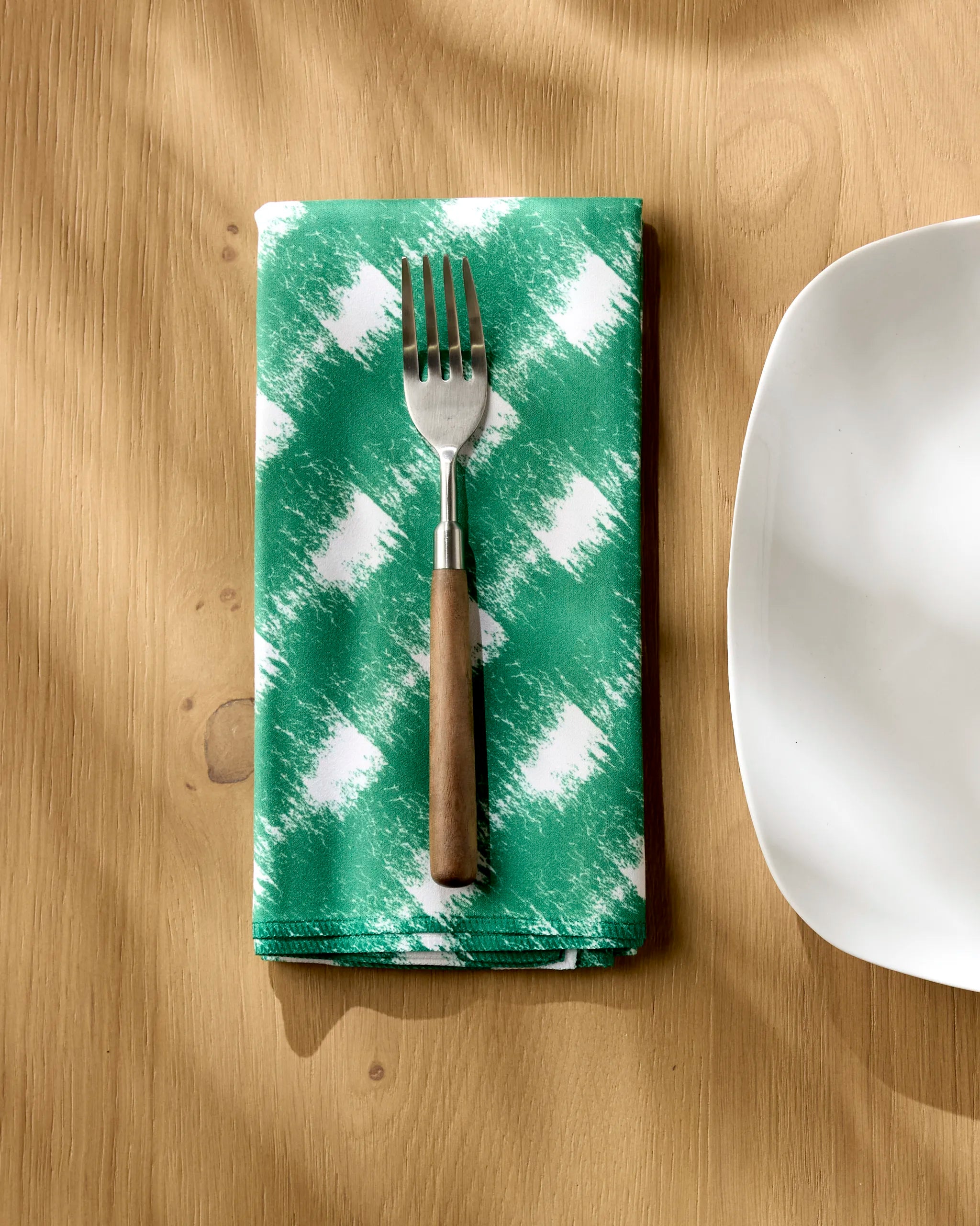 A fork with a wooden handle rests on Geometry’s Hairy Check Green absorbent cloth napkin next to a white, square plate on a light wooden table.