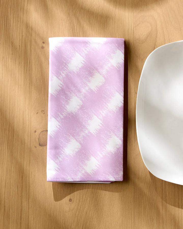 A folded Geometry Hairy Check Pink napkin from the Dinner Napkin Set rests on a light wooden table beside the edge of a plain white plate.