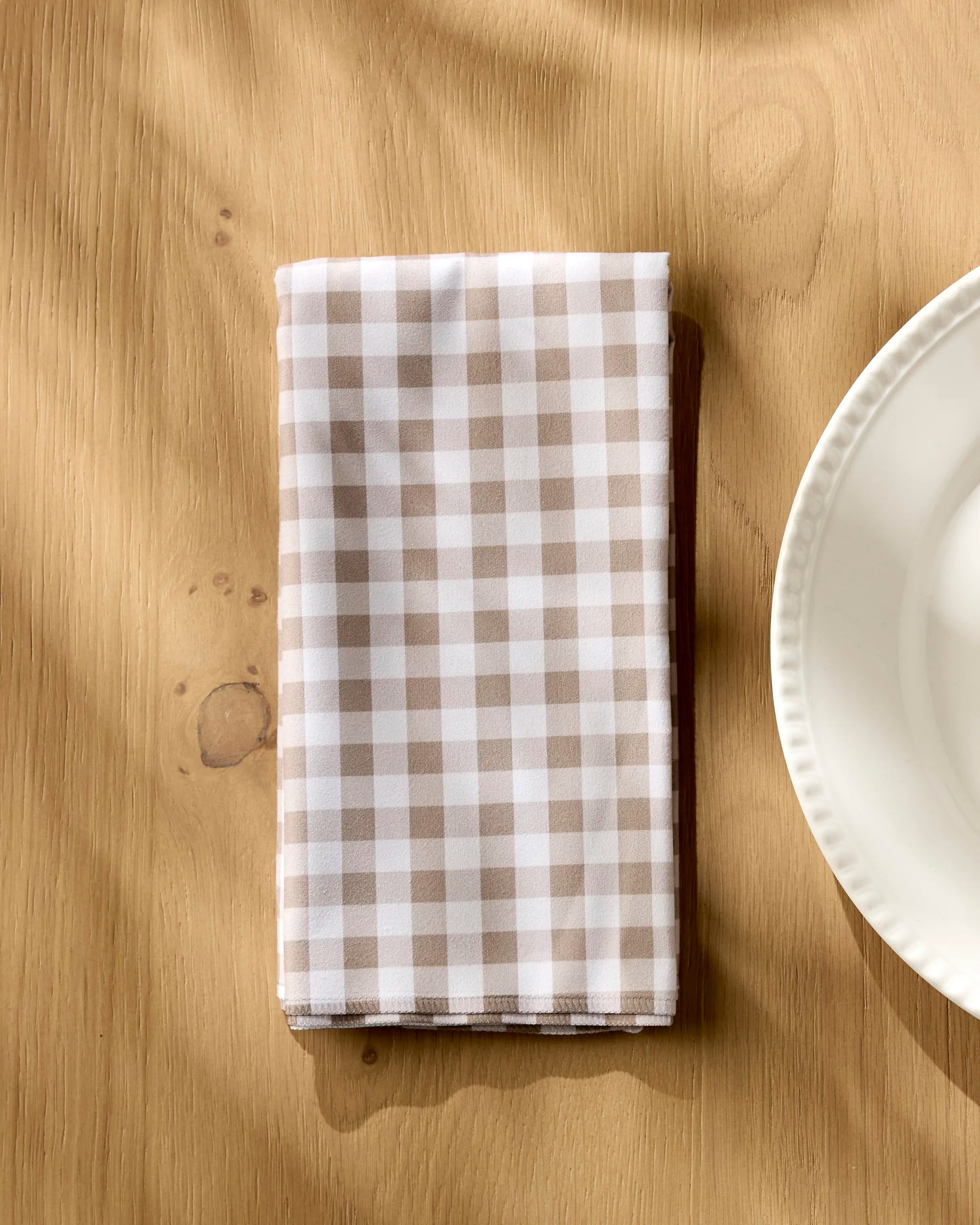 A neatly folded Hazel Gingham napkin by Geometry rests on a light wooden table beside a white ceramic plate, with sunlight casting gentle shadows over the scene.