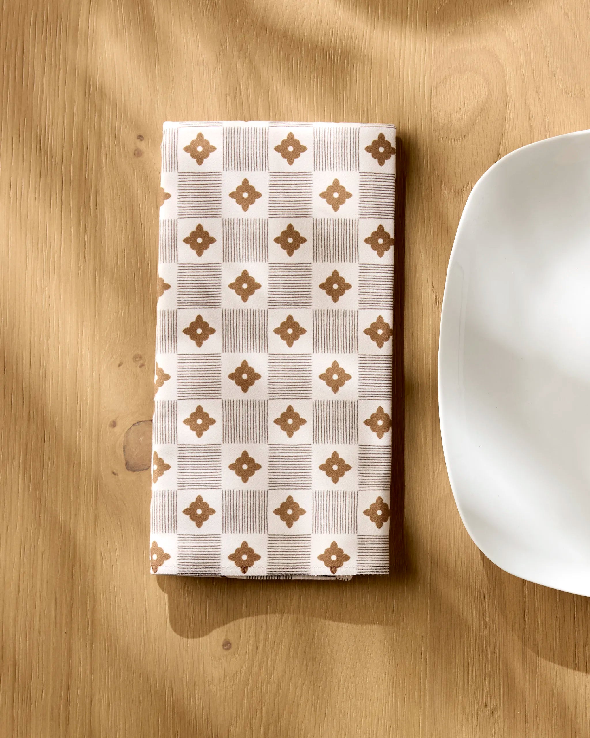 A folded beige Heritage Stamp dinner napkin from Geometry, crafted from Geosuede fabric, sits on a wooden table next to a white, square-edged plate as sunlight casts soft shadows across the scene.