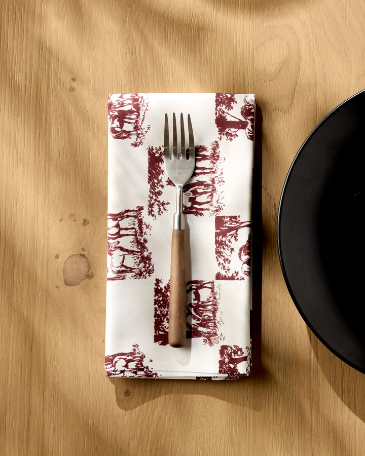 The Geometry Horse Check Cream recycled dinner napkin is folded under a fork with a wooden handle, next to the edge of a black plate on a light wooden table.