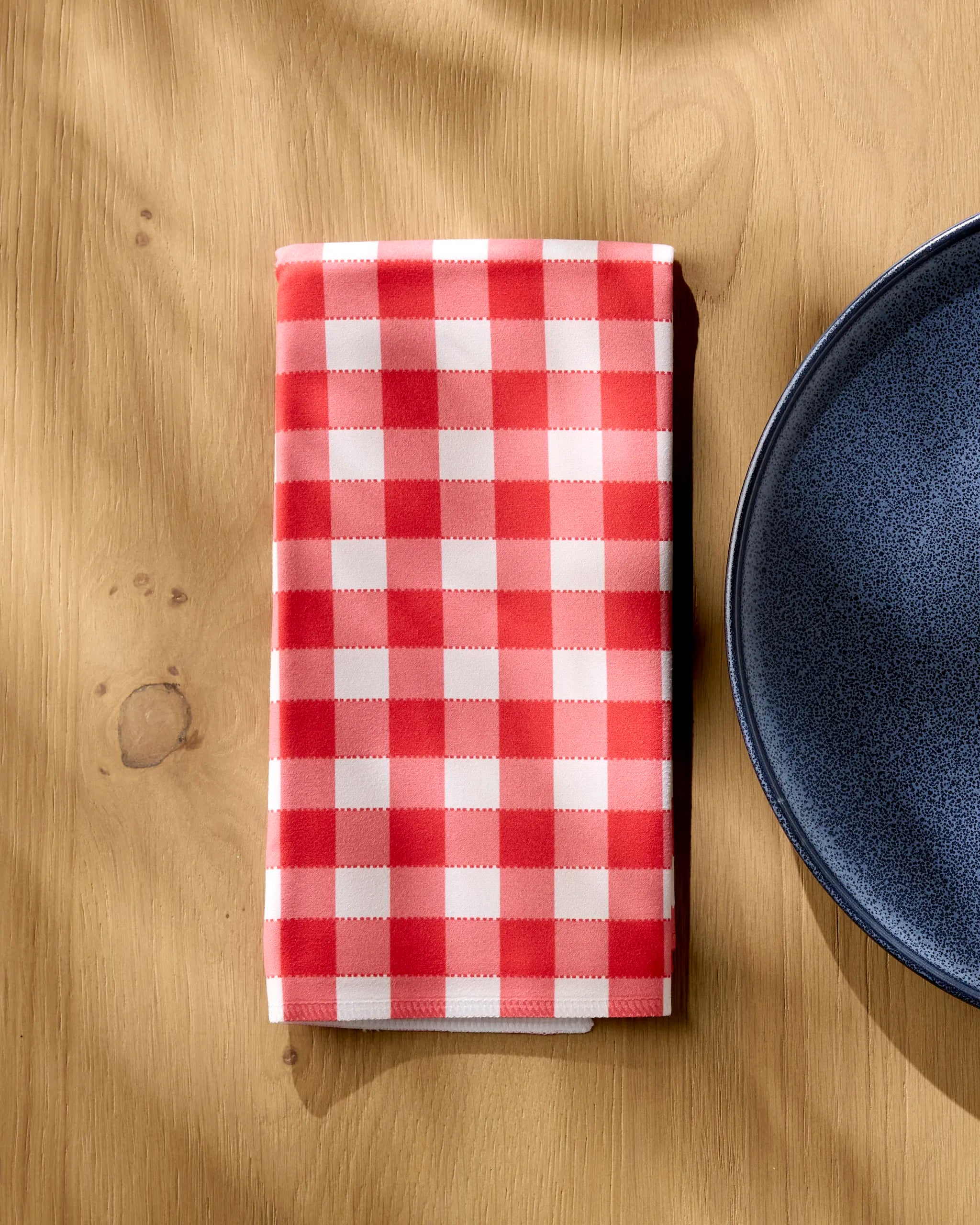 A Geometry Ripe Tomato Gingham recycled dinner napkin is neatly folded on a wooden table next to the edge of a dark blue ceramic plate.