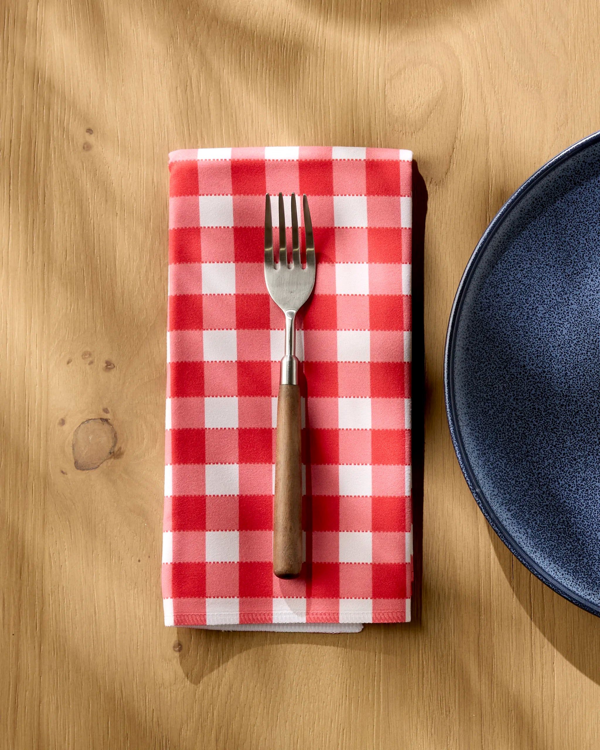 A fork with a wooden handle rests on a Geometry Ripe Tomato Gingham absorbent cloth napkin, placed on a wooden table next to a dark blue ceramic plate.