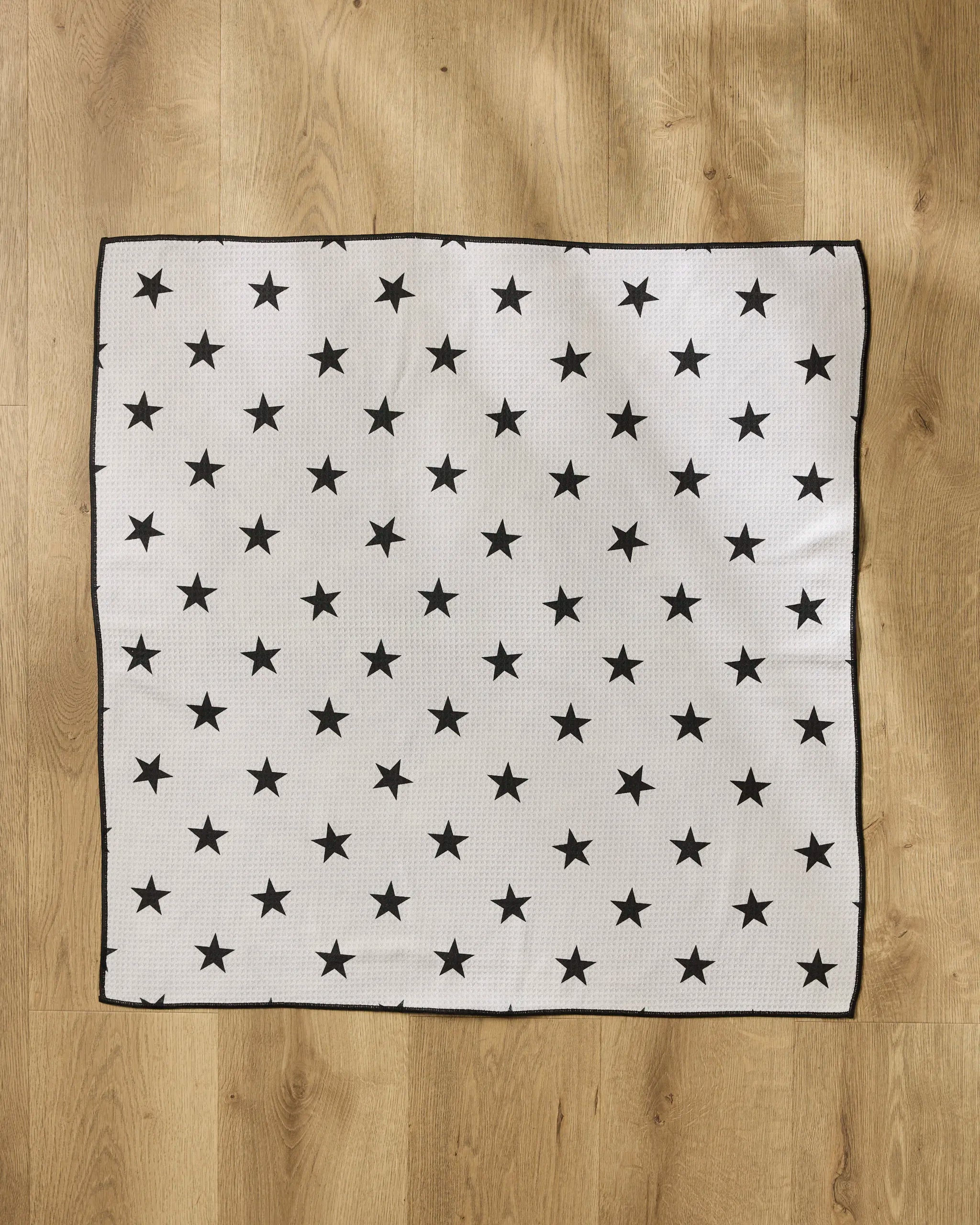 The Geometry Bark dog towel features a white base with evenly spaced black stars and black-stitched edges, offering both absorbency and style for your pet. Laid flat on a wooden floor, it blends practicality with modern design.