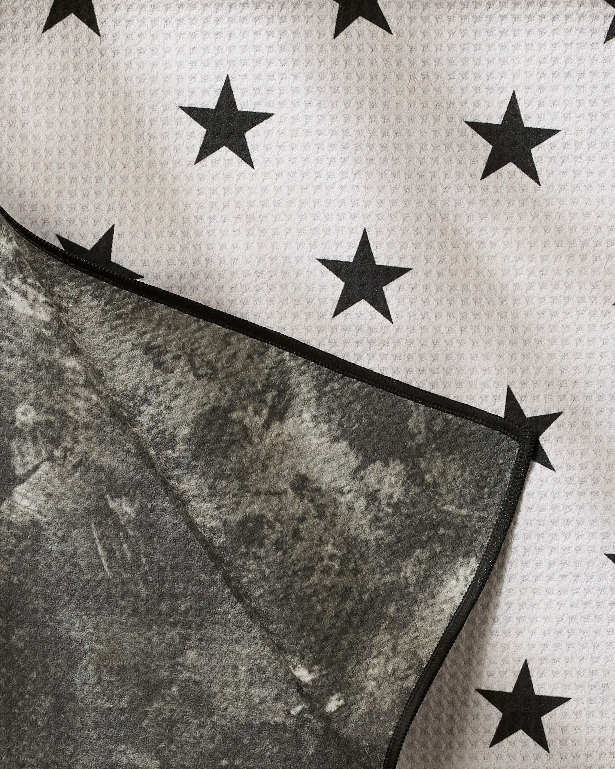 A close-up of Geometry's Bark dog towel shows its absorbent white surface with black stars, a folded corner revealing the mottled gray underside and black edge.