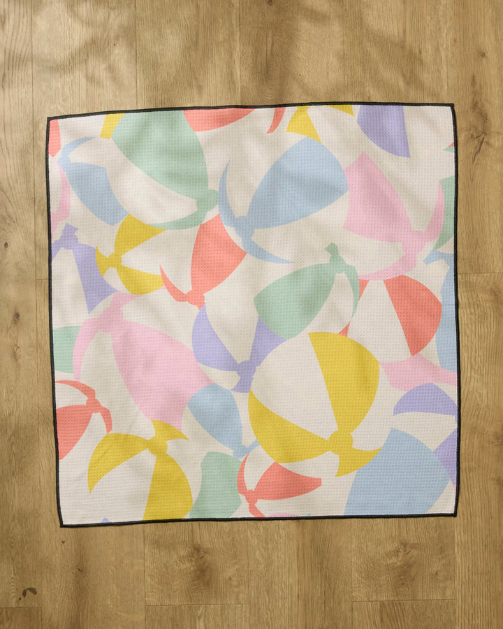 The Geometry Beach Ball dog towel is a square, absorbent towel featuring a colorful pattern of pastel beach balls outlined in black, shown flat on a wooden floor.