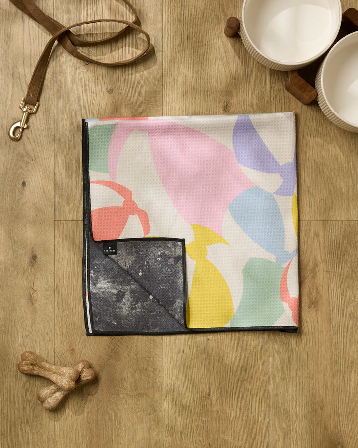 A Geometry Beach Ball dog towel with colorful abstract shapes lies on a wooden floor beside two white bowls on a wooden stand, a leather leash, and two dog bones.