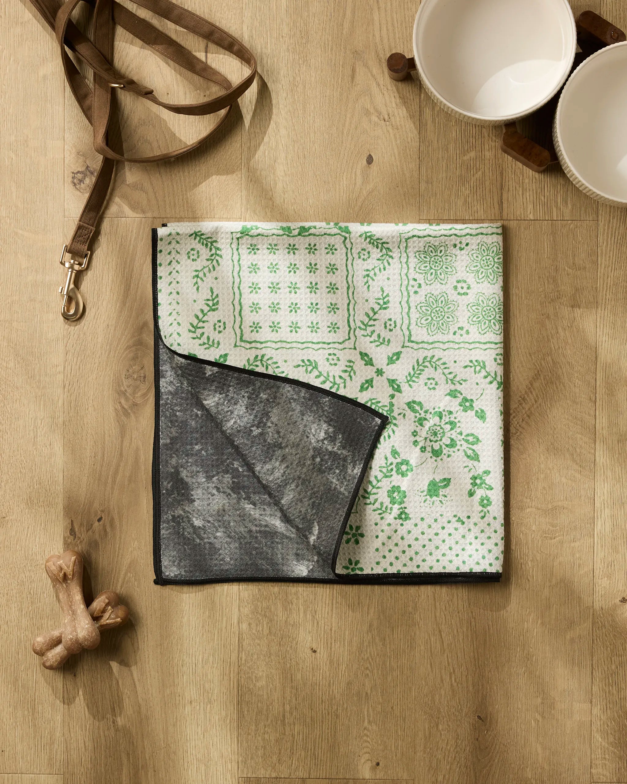 The Geometry Blossoming Bandanna Green pet mat, crafted from recycled materials and featuring a green and white pattern, is shown with a flipped corner on a wooden floor beside a leash, two bowls on a stand, and two dog bones.