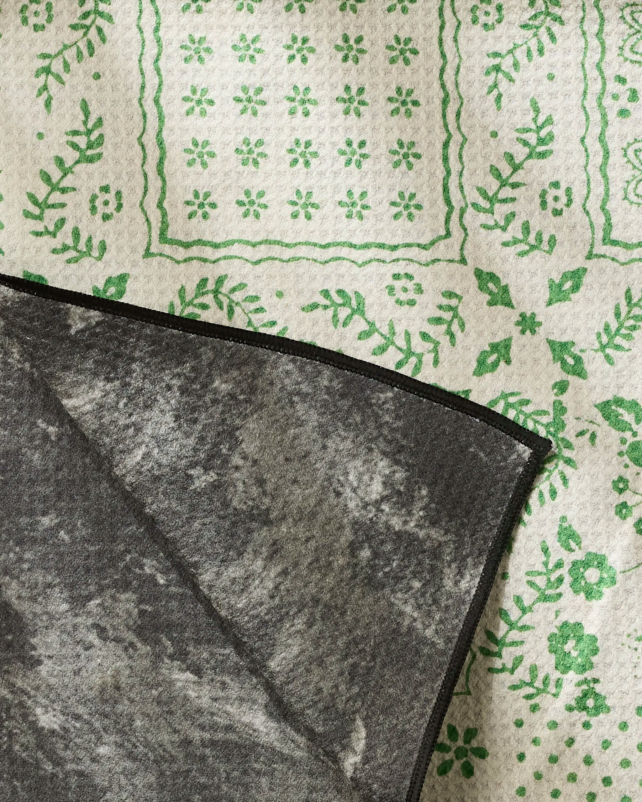 The Geometry Blossoming Bandanna Green rug features a cream base with green floral and grid designs, partially flipped to show its dark gray, textured backing crafted from recycled materials.