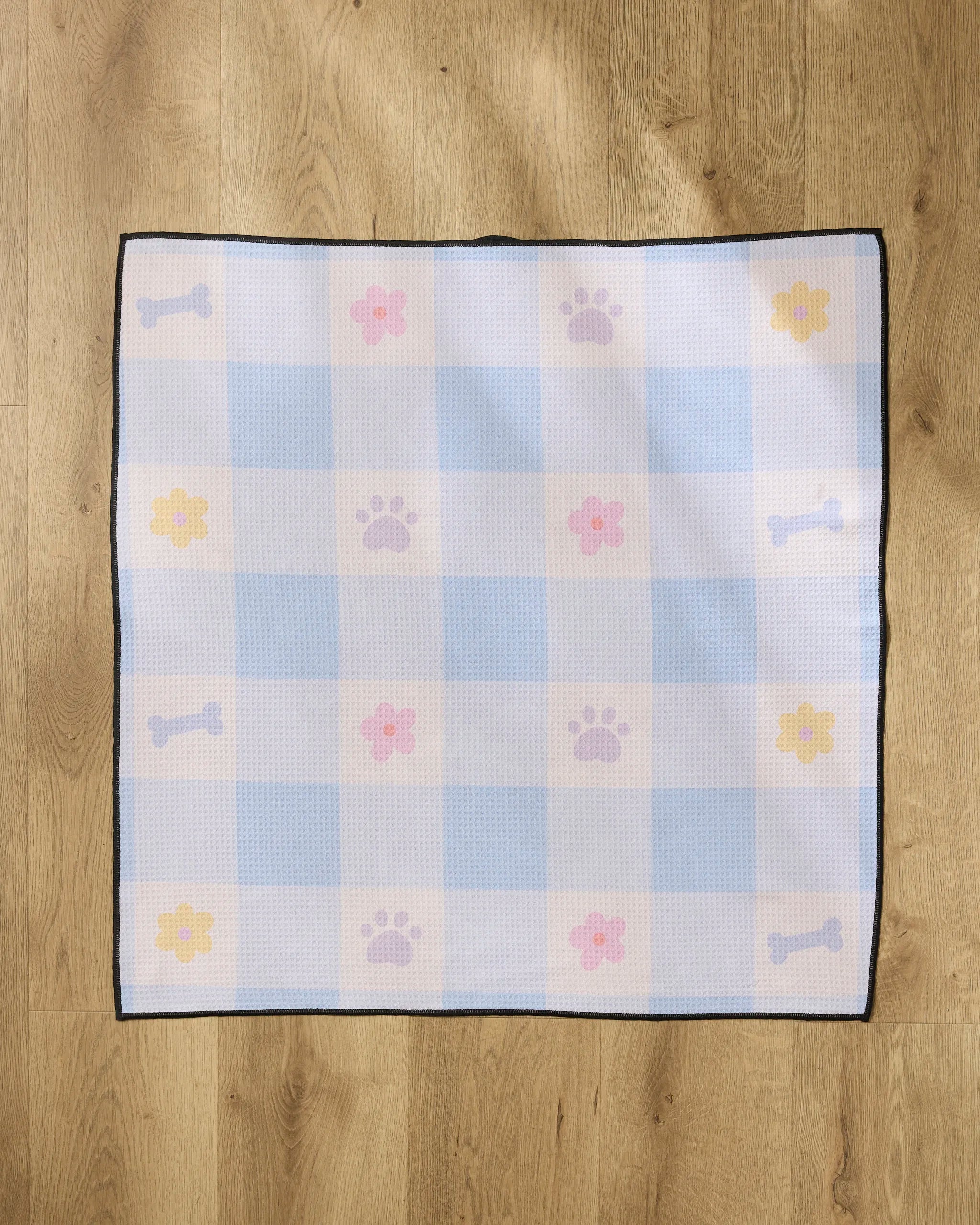 The "Blue Sky Picnic Dreams" towel by Letters By Shells is a square, highly absorbent, recycled dog towel with blue and white checks, purple paw prints, yellow and pink flowers, and blue dog bones.