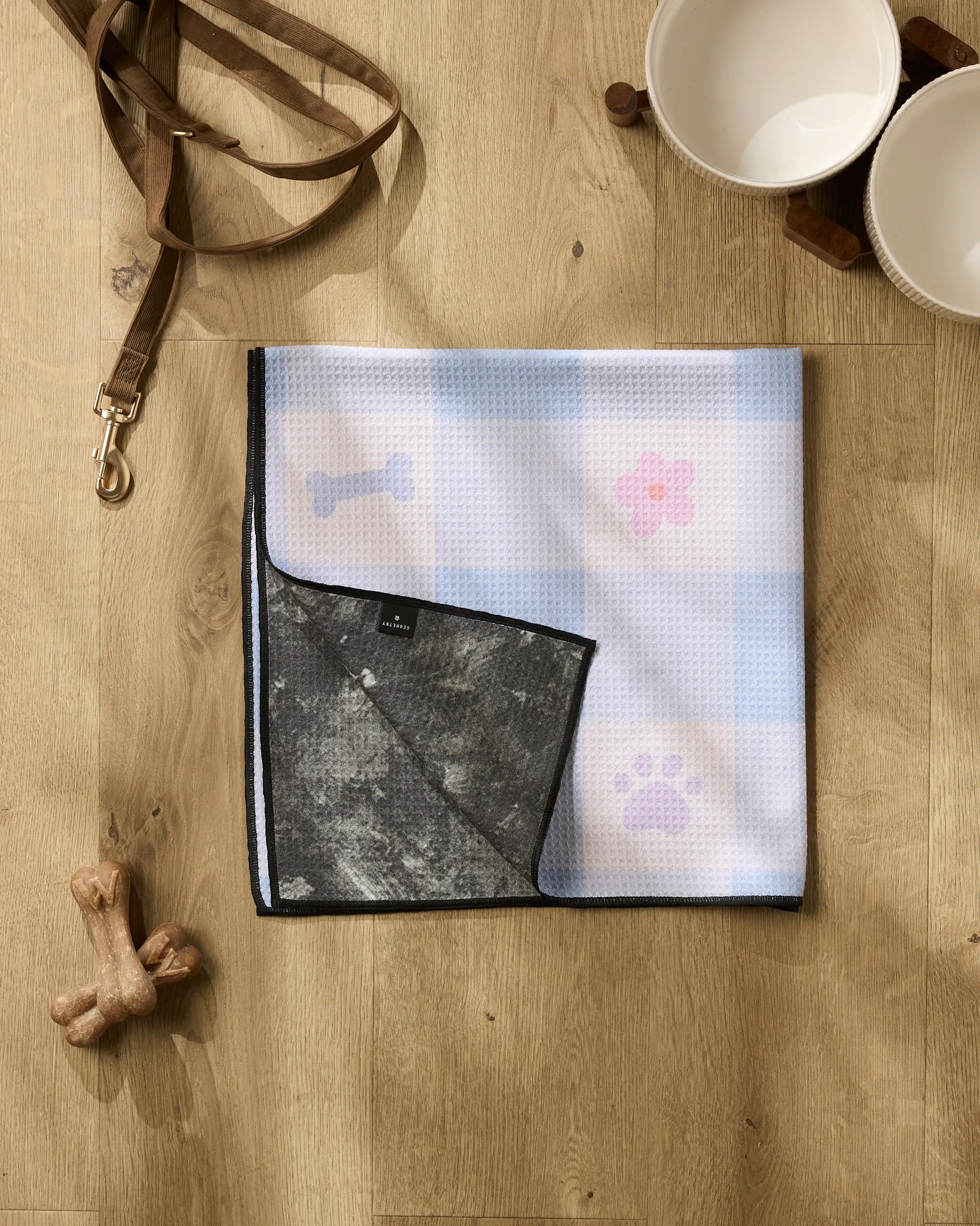 The Blue Sky Picnic Dreams set by Letters By Shells features a dog leash, two empty bowls on a stand, a bone-shaped treat, and an absorbent towel with paw and bone prints arranged on a wooden floor.