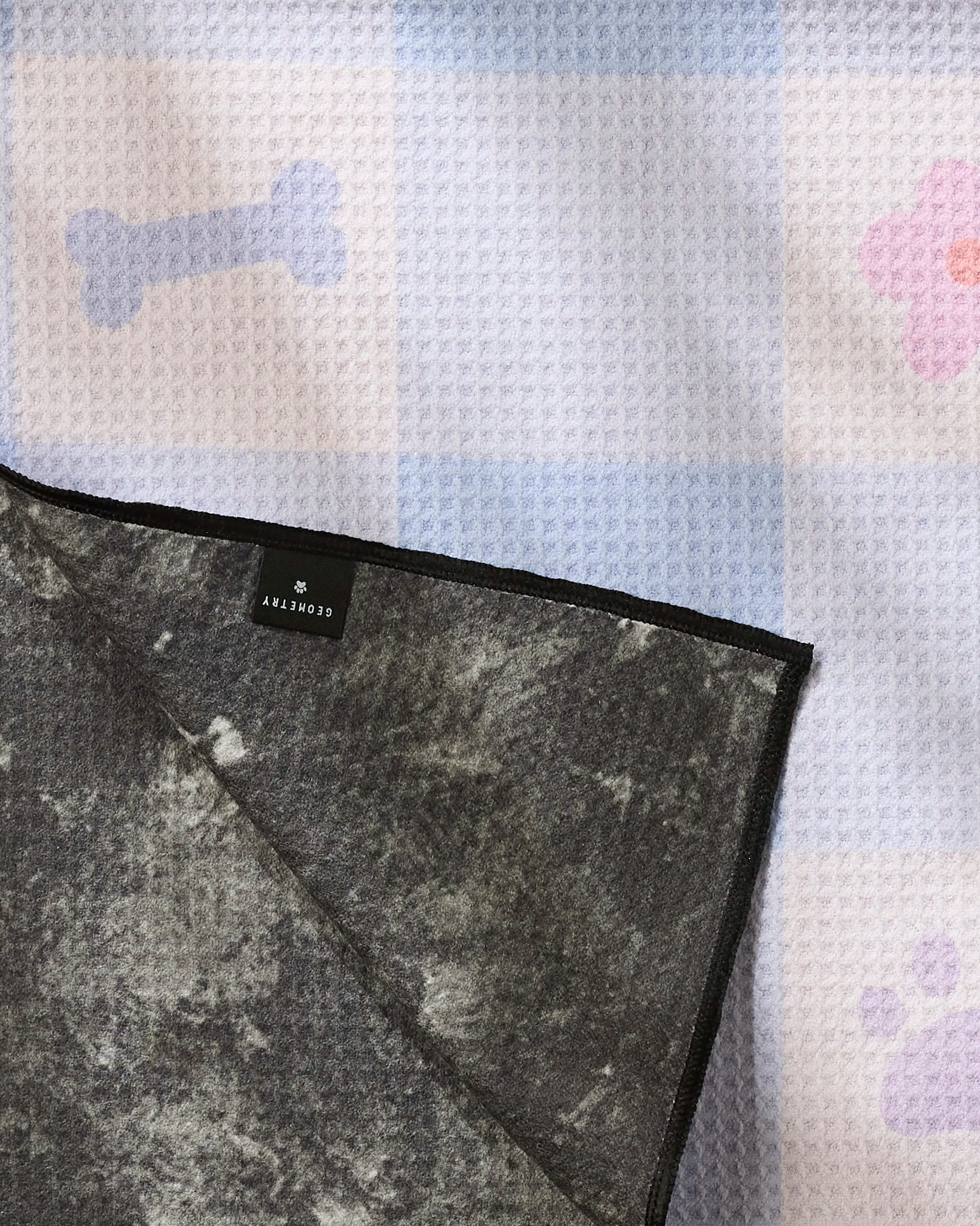 A close-up of the Blue Sky Picnic Dreams pet mat by Letters By Shells features a blue and white checkered pattern with bone and paw prints. Made from recycled materials, its corner folds over to reveal a dark gray underside with a black label.