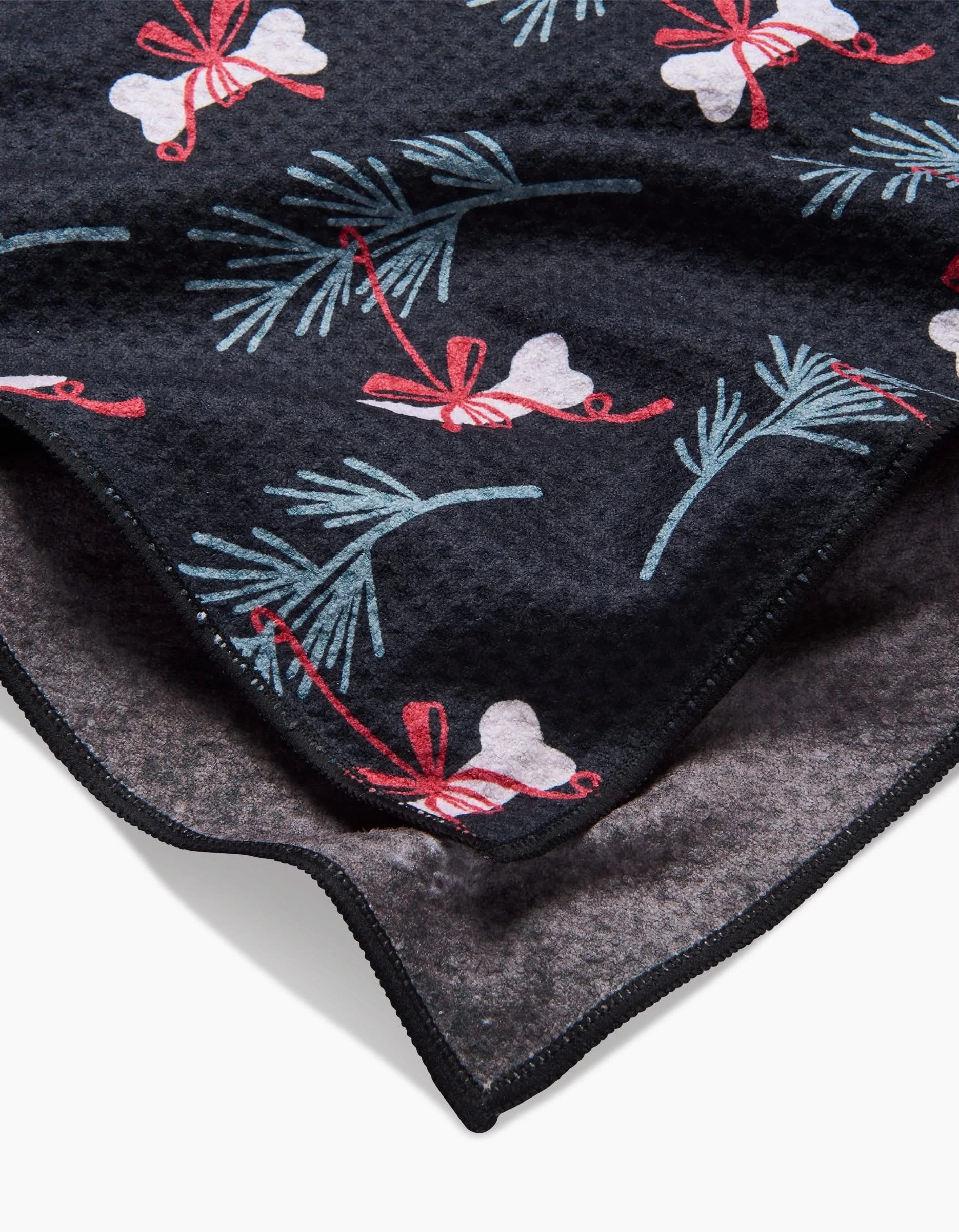 A close-up of the Bones For Christmas dog towel by Charlie Rowan Designs shows its dark textured, recycled fabric with white bells, red bows, and green pine branches, folded to display the absorbent front and back.