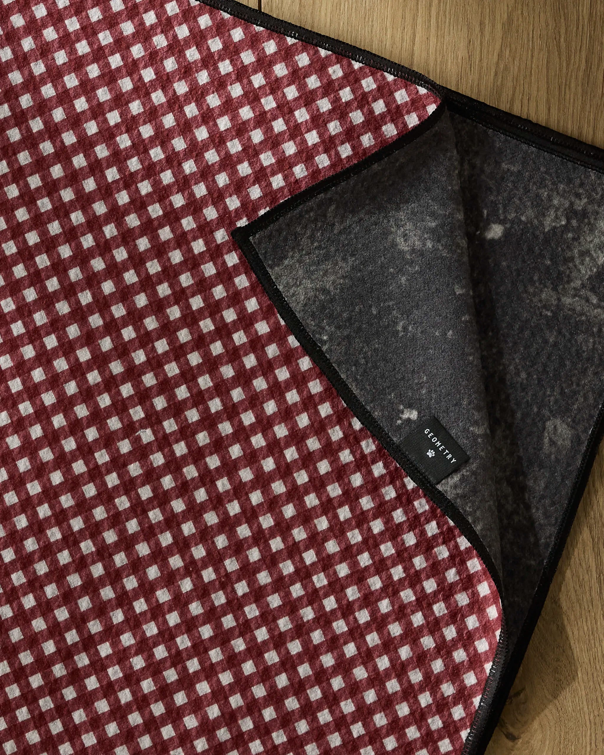 The Bramble Red dog towel by Geometry features a red and white checkered pattern with a dark gray underside and a small black "COTTON 60%" label. Made from recycled materials, it rests on a wooden surface.