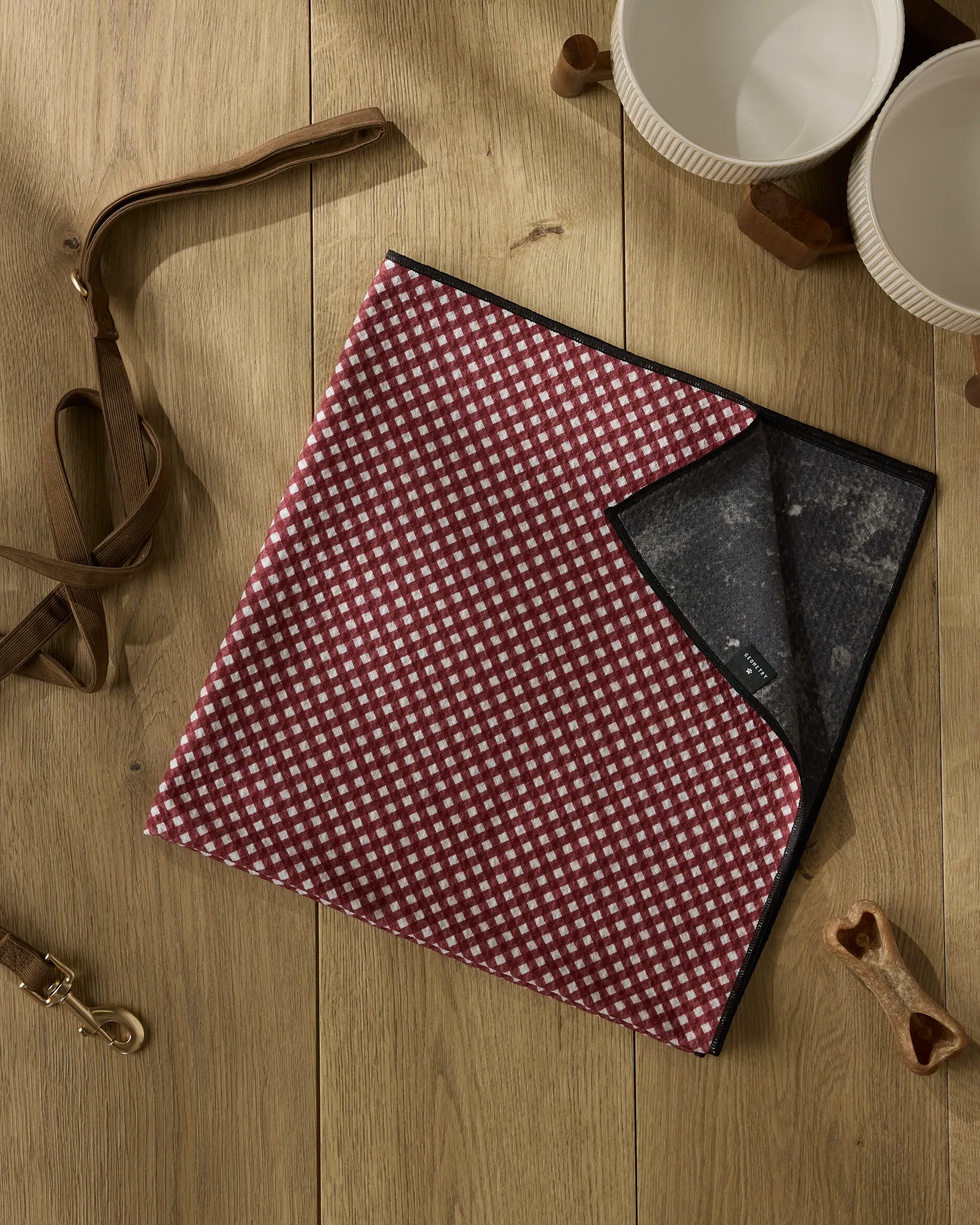 The Geometry Bramble Red towel, featuring a red and white checkered pattern, black edge, and high absorbency, is partially folded on a wooden floor. Nearby are a brown leather leash, treats, and two white bowls.