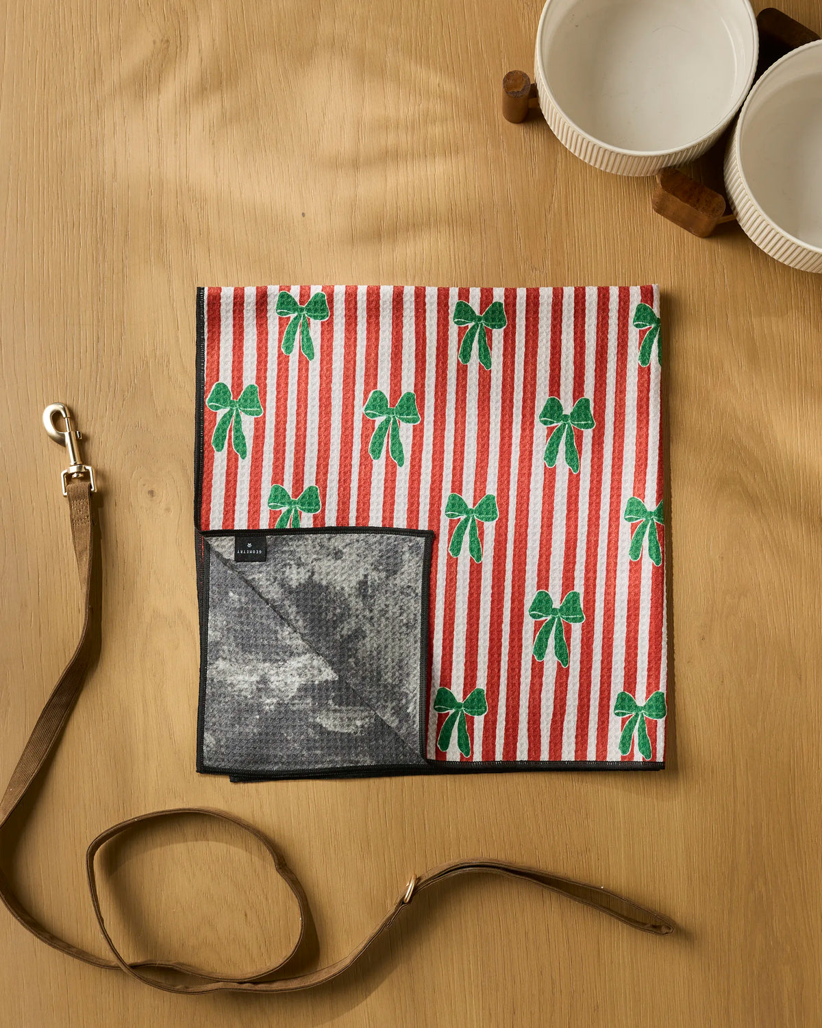 A Geometry Candy Bows pet mat, featuring red and white stripes, crafted from recycled materials and decorated with green bows, rests on a wooden table. Nearby are a highly absorbent dog towel, a brown leash, and two white ceramic pet bowls.