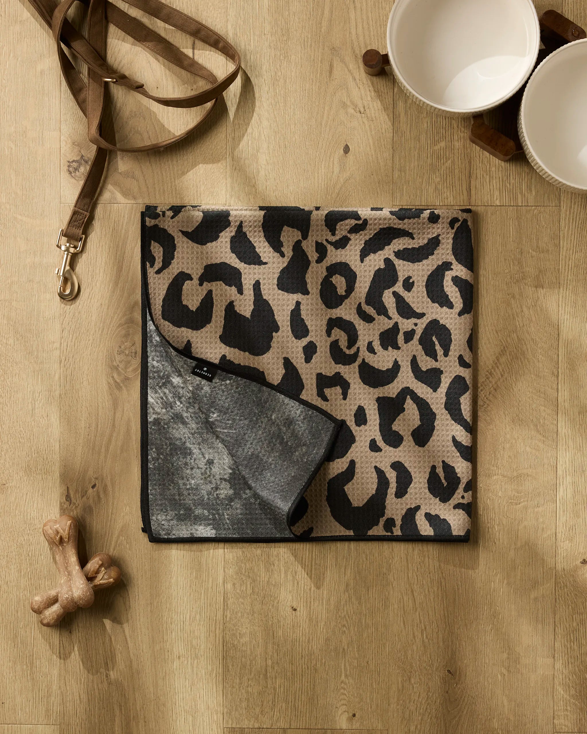 The Cheetah Hound dog towel by Geometry, made from recycled materials, lies folded on a wooden floor beside a brown leash, two ceramic pet bowls on a wooden stand, and two chew bones.