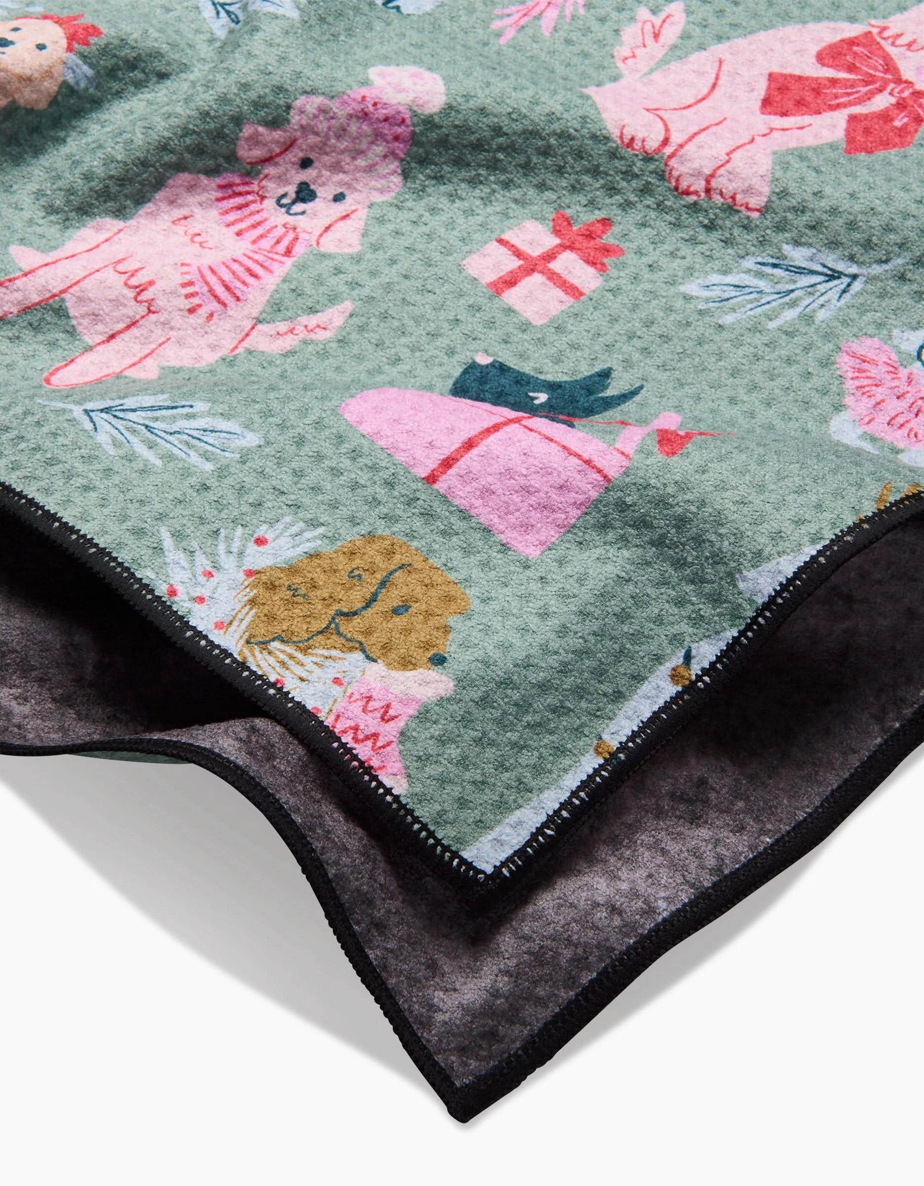 A close-up of the Christmas Doggos towel by Janna Sue Design, made from recycled materials with a playful dog, gift, and pine pattern on green, dark edging, and a soft, dark inner lining.