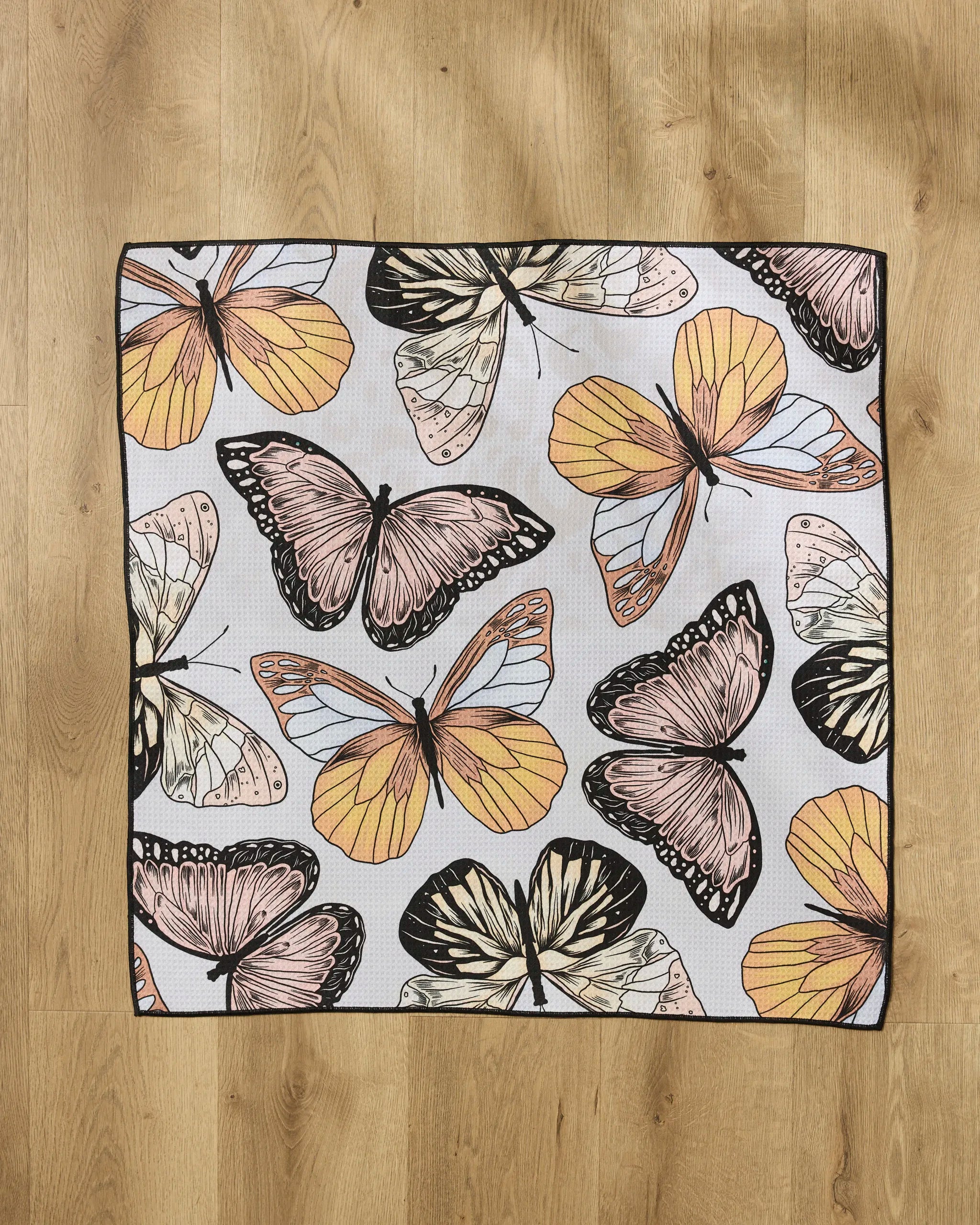 The Geometry "Cute Butterflies" square dog towel is made from recycled materials and displays large, vibrant butterflies in orange, yellow, pink, and black on a light wood floor.
