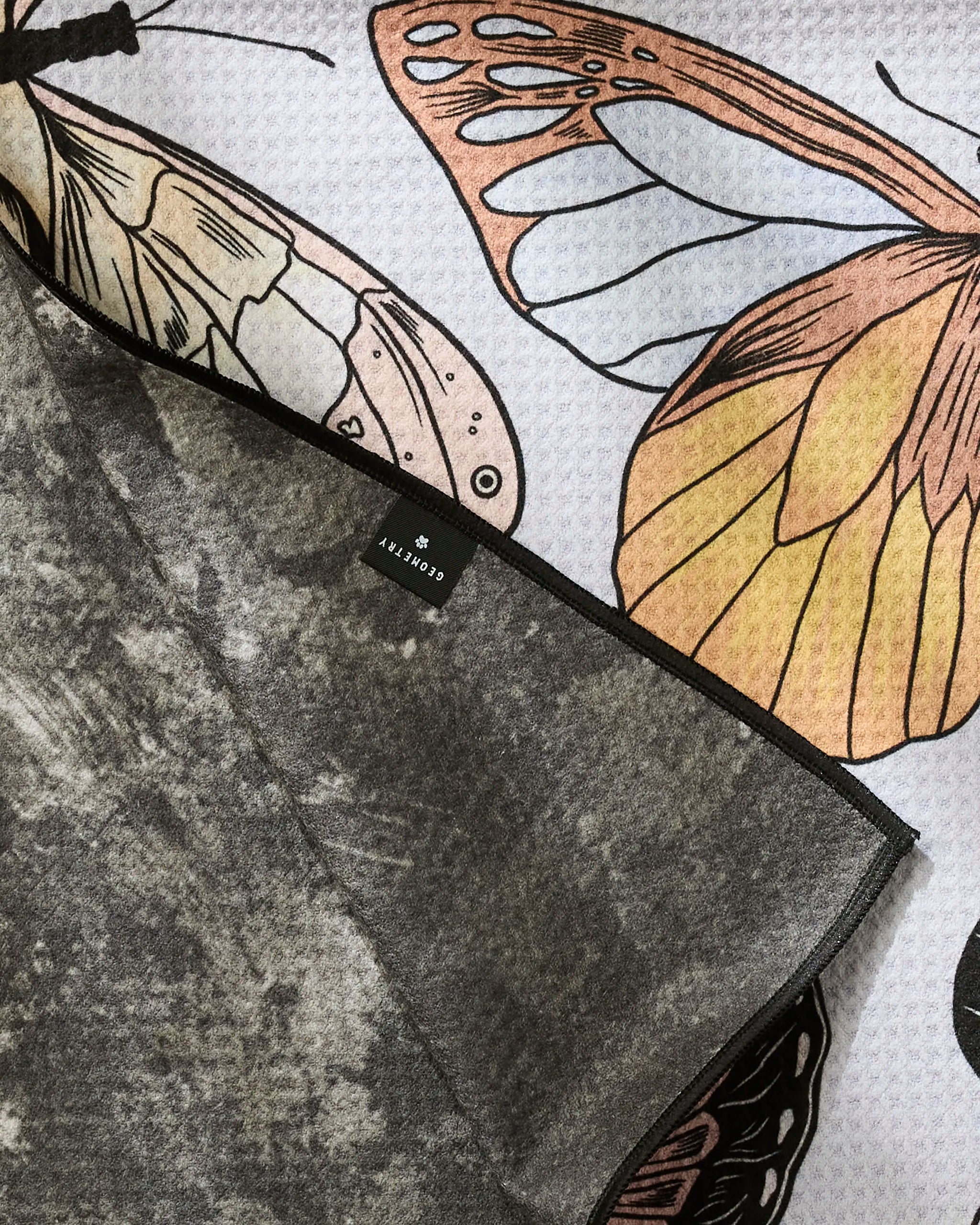 A close-up of the Geometry "Cute Butterflies" dog towel, featuring butterfly-patterned fabric with a textured gray underside and a small black brand tag.