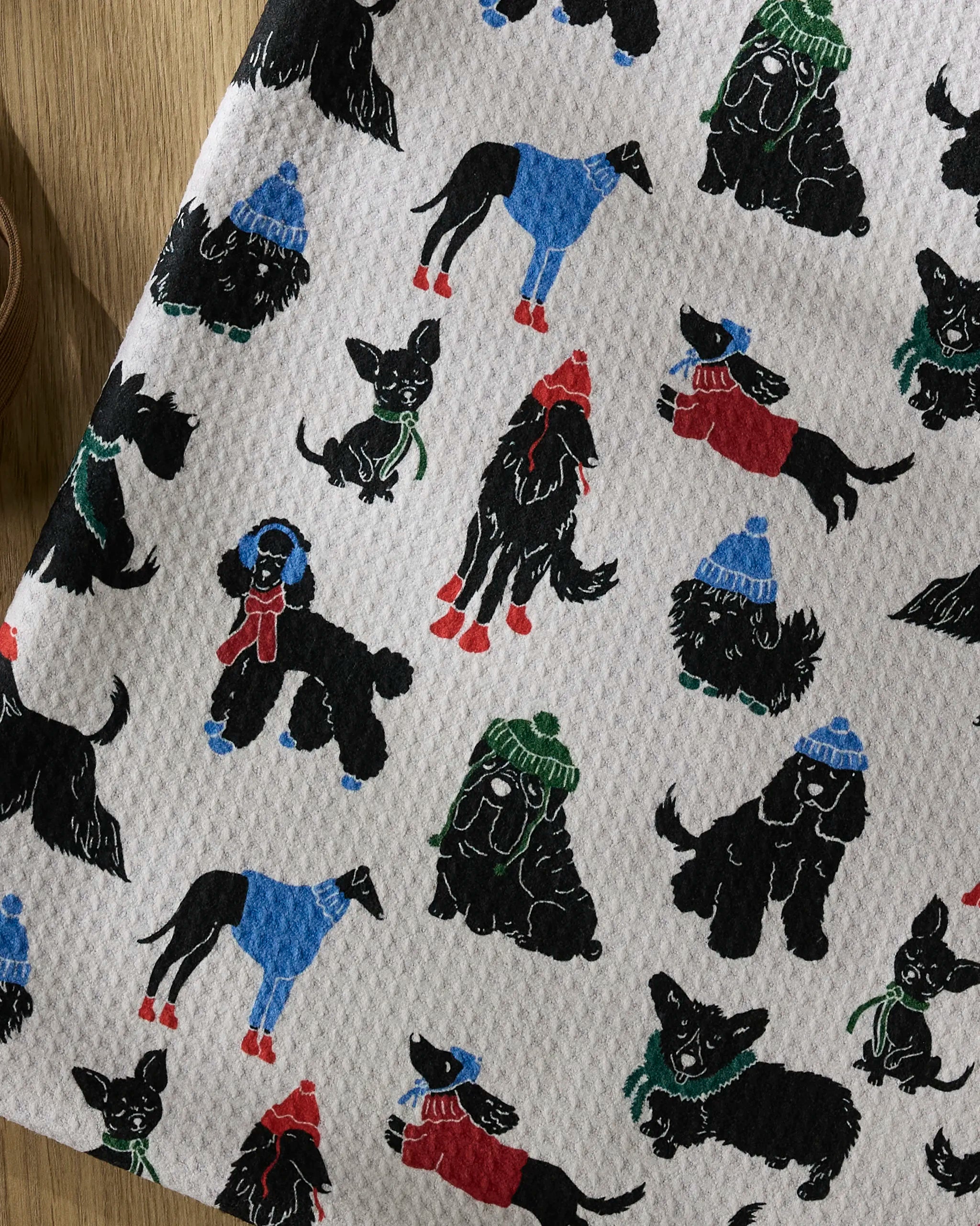 The Geometry Dogue Magazine dog towel is highly absorbent, made from recycled materials, and features black dogs in colorful winter hats, scarves, and sweaters on a white background.