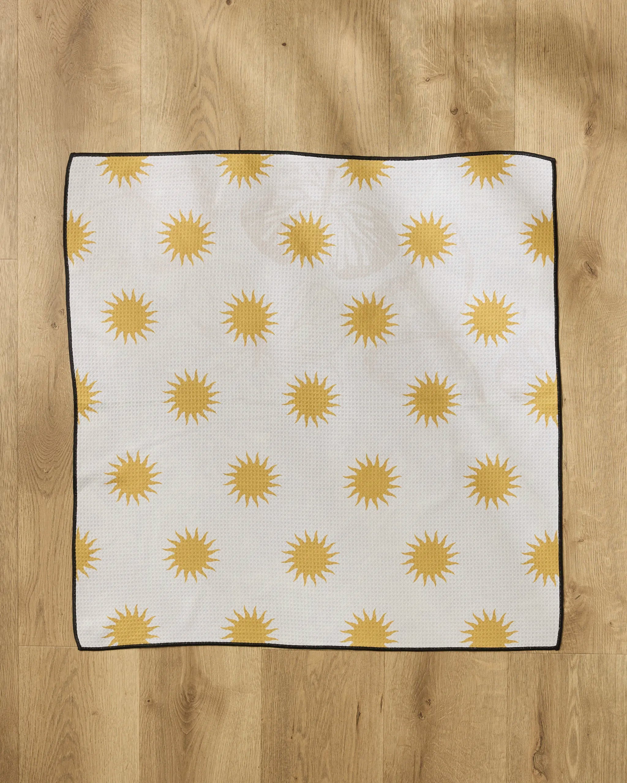 The Geometry Feeling Sunny dog towel, a square white towel made from recycled materials with yellow sun shapes and a black border, lies flat on a wooden floor.