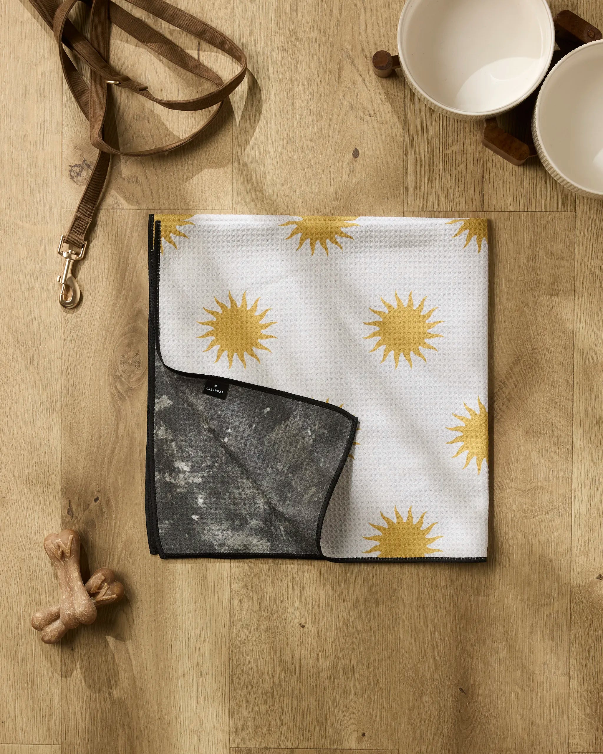 The Geometry “Feeling Sunny” towel, highly absorbent with yellow sun patterns, lies partly folded on a wooden floor. Nearby are a brown leash, two ceramic bowls, and two wooden dog bones.