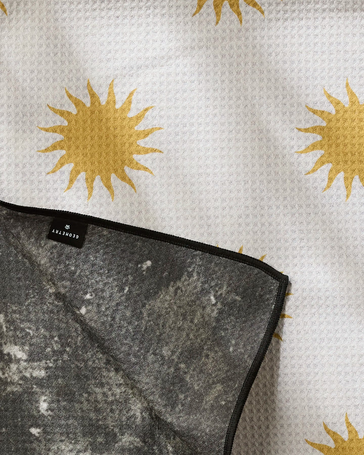 The Feeling Sunny blanket by Geometry features textured white fabric with yellow sun patterns, a partially folded back dark gray underside crafted from recycled materials, and a label bearing the brand name.