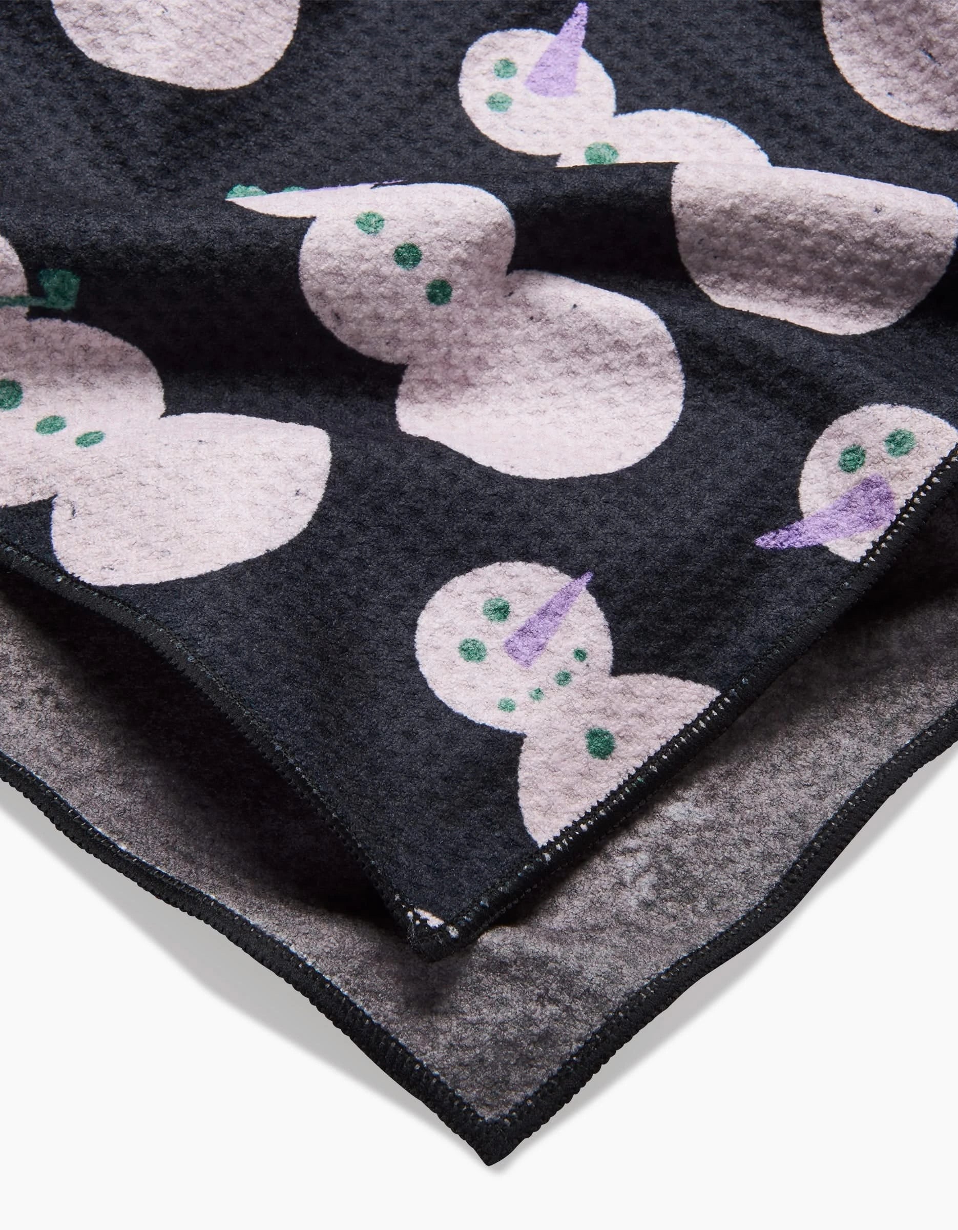 A close-up of the Felicity Black towel by Amy MacCready shows its textured dark fabric with light snowmen featuring green buttons and purple or green carrot noses. One corner is folded, revealing the reverse side.