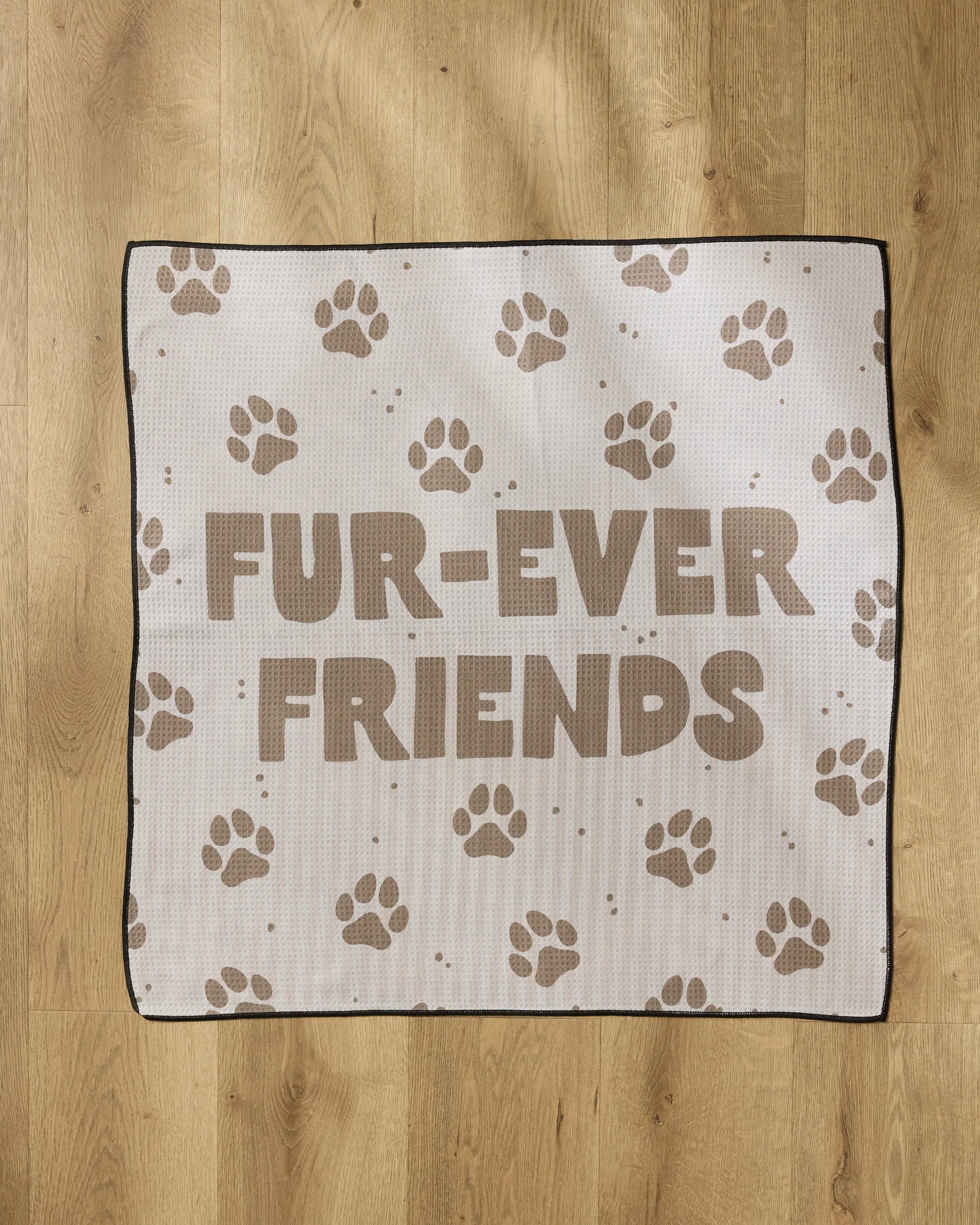 The Geometry Fur Ever Friends mat features brown paw prints and bold "FUR-EVER FRIENDS" text on an absorbent white surface, displayed on a wooden floor.