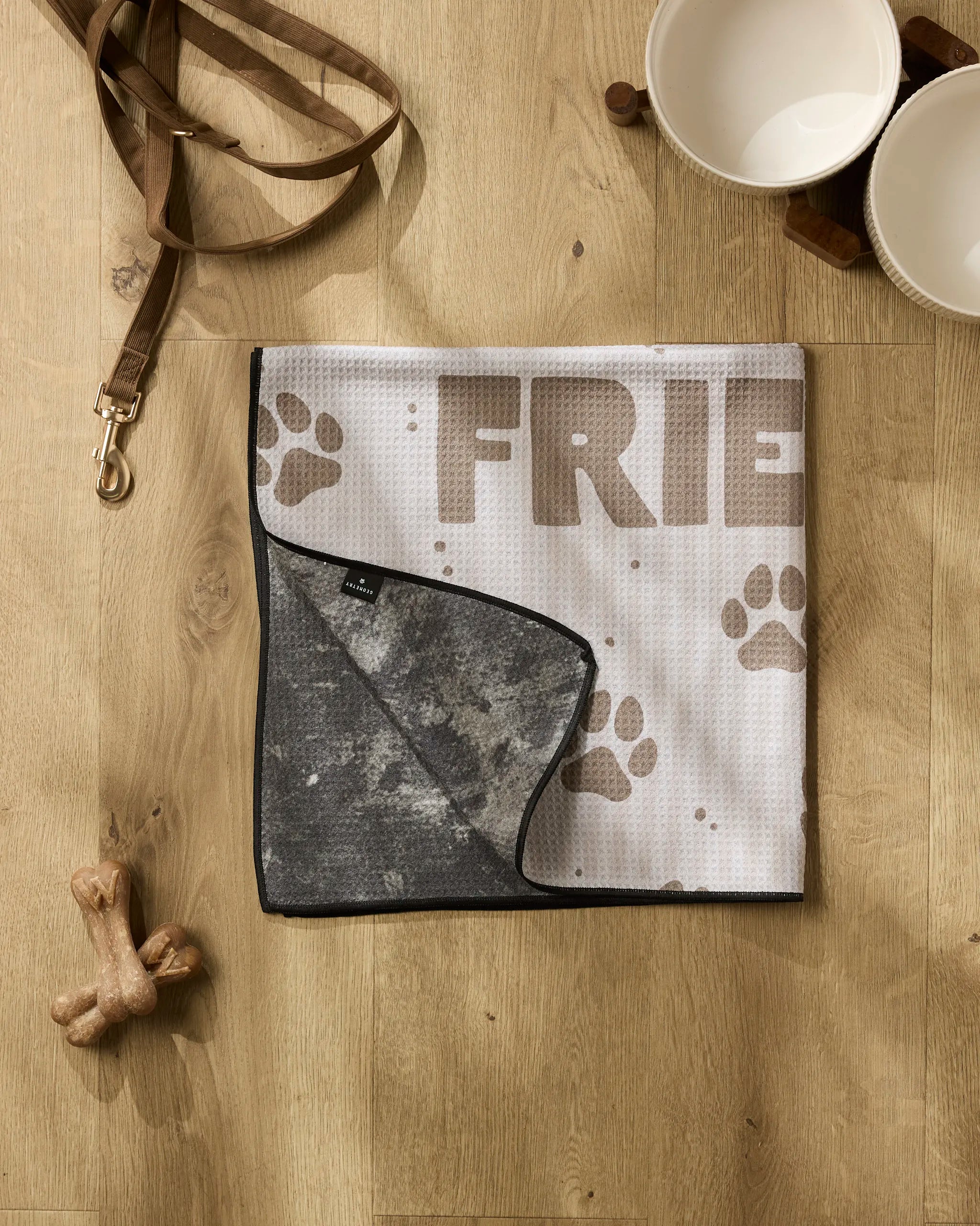 Geometry's Fur Ever Friends set, including a dog leash, two white pet bowls, a super absorbent towel, bone-shaped chew toy, and a paw print FRIEND pet mat, is arranged on a wooden floor.