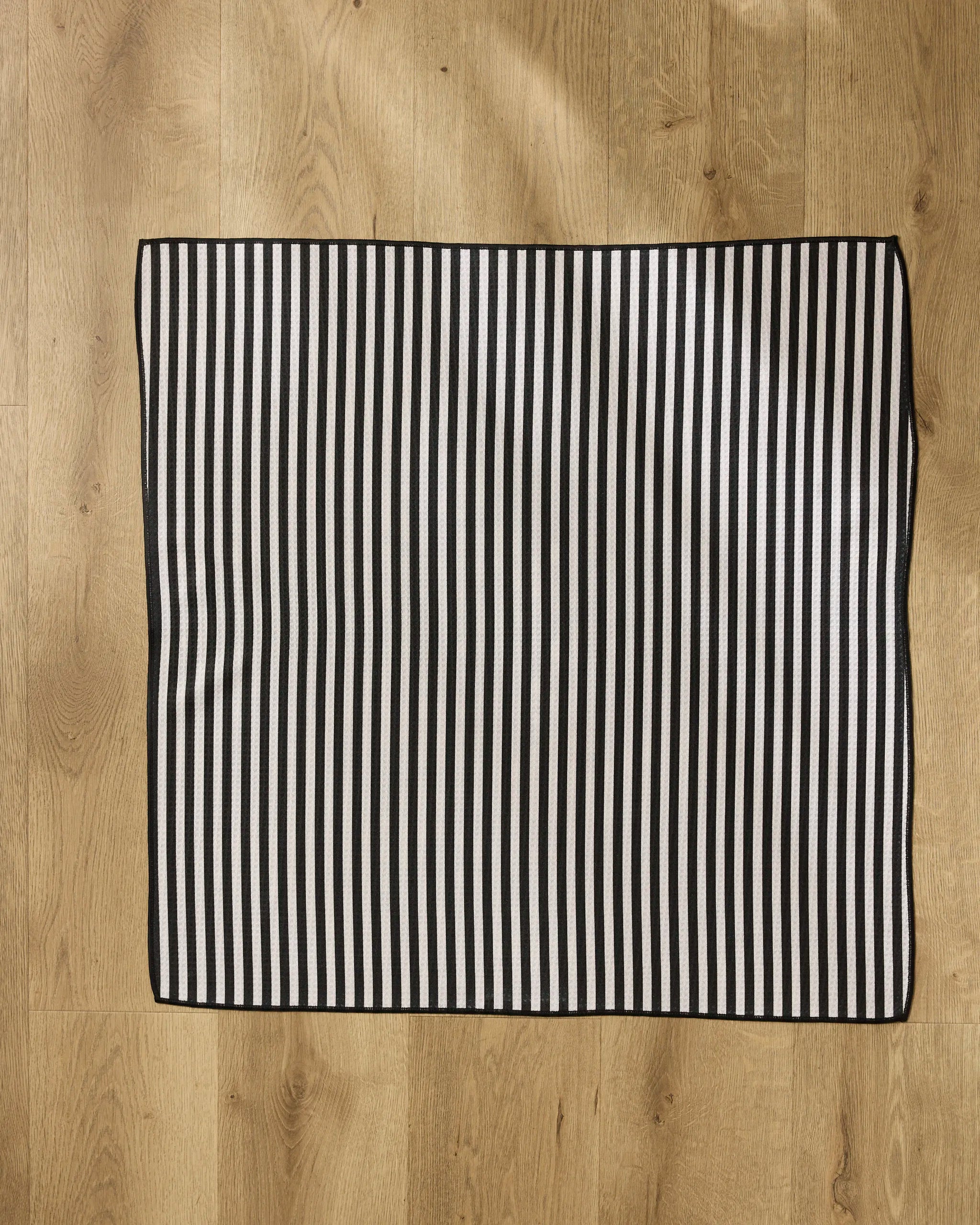 The Geometry Good Pup mat, a rectangular and highly absorbent piece with black and white vertical stripes, rests on a light wooden floor illuminated by natural light.