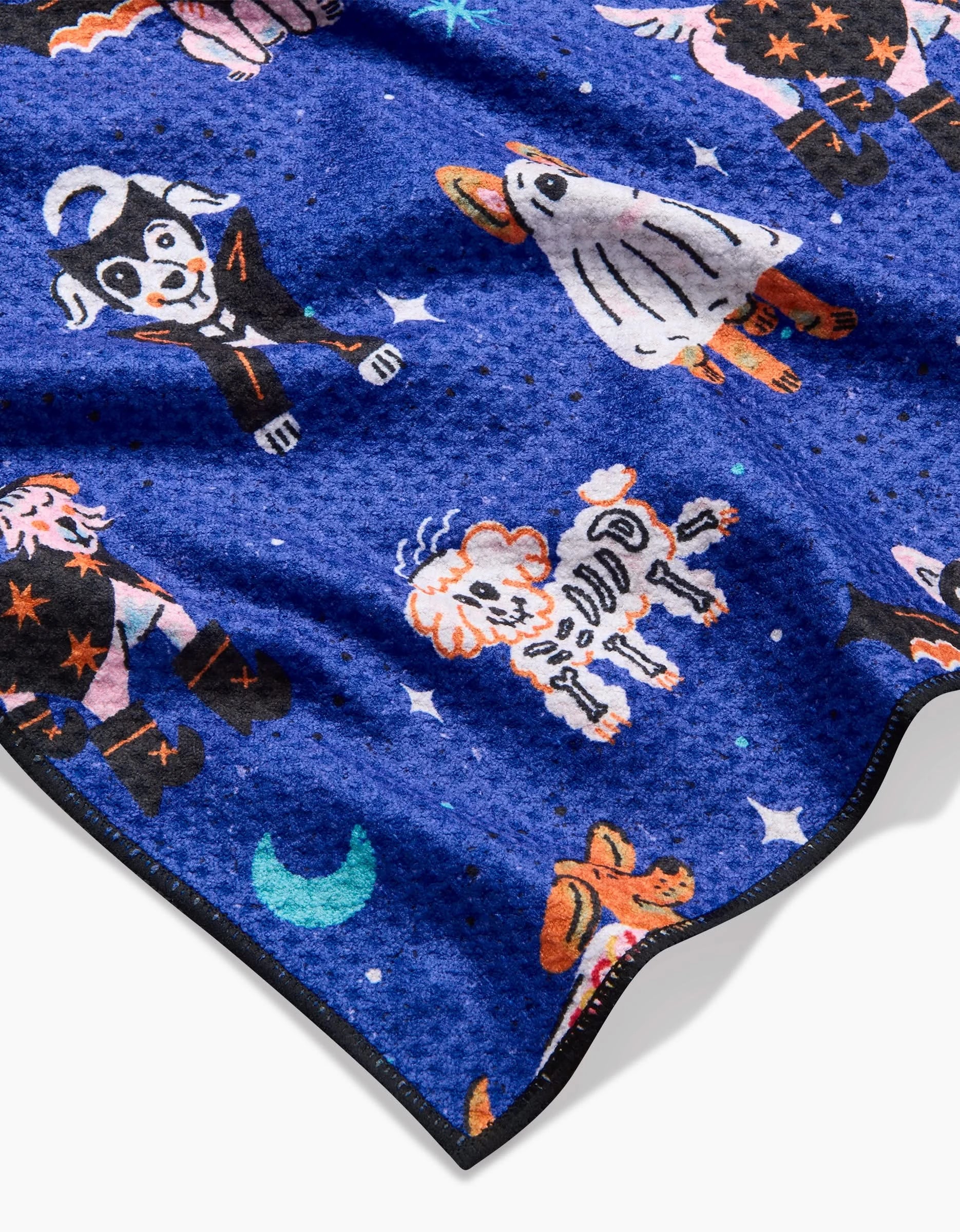 The Halloween Hounds dog towel by Camila Leão is blue, textured, and adorned with festive Halloween designs—costumed dogs, ghosts, skeletons, and stars. Made from highly absorbent recycled materials for both fun and practicality.