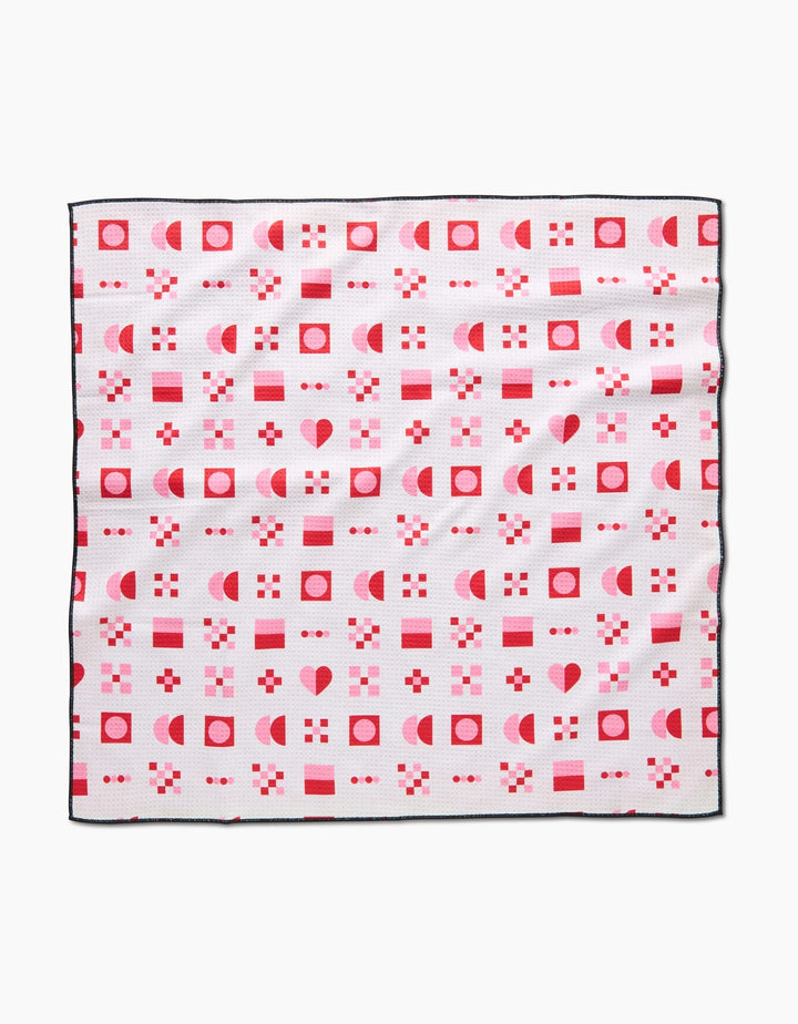 The Laura Supnik Heart Quilt is a square absorbent dog towel with a white background, black edges, and a grid pattern of red and pink hearts, squares, circles, and geometric shapes.