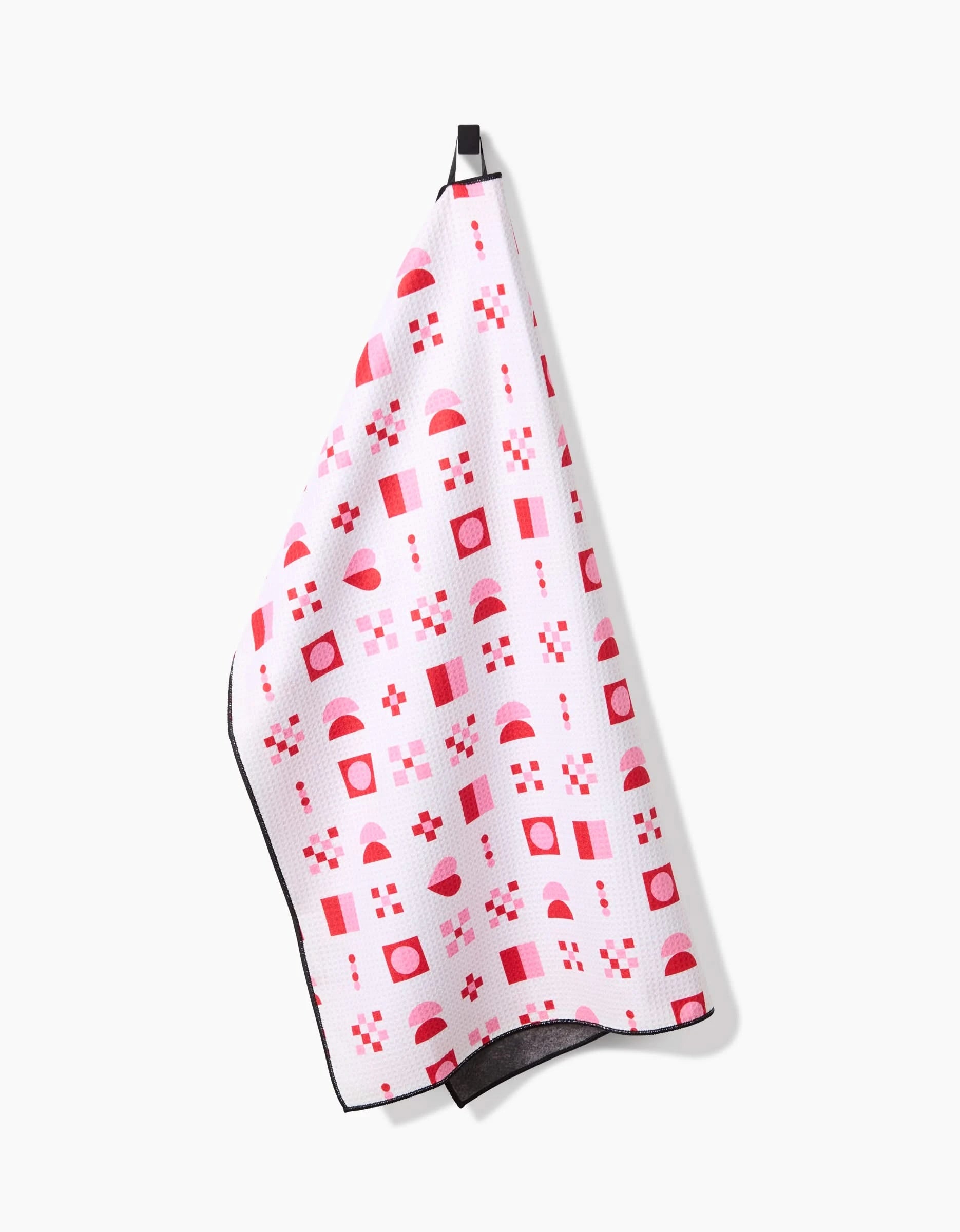 The Heart Quilt towel by Laura Supnik, featuring red and pink geometric shapes on white, hangs on a hook against a plain white background.