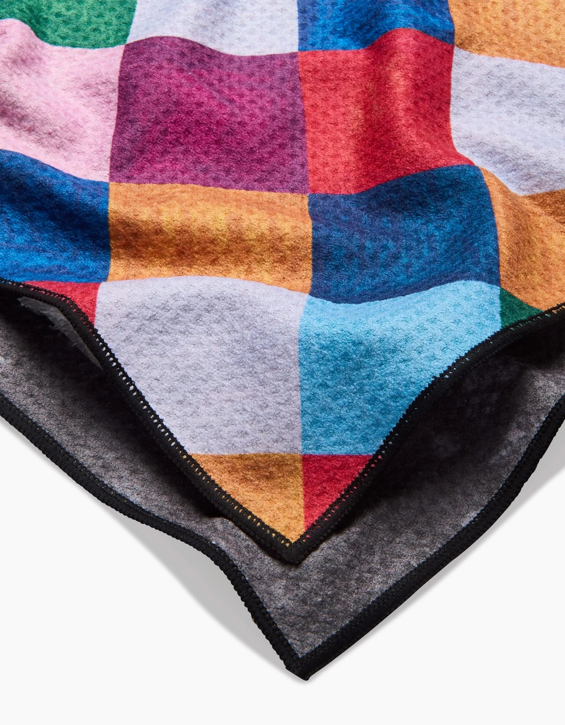 The Holiday Harlequin dog towel by Daria Solak features a soft, textured design with a vibrant patchwork of red, blue, yellow, orange, and pink squares—crafted from recycled materials and shown over a dark gray backing.