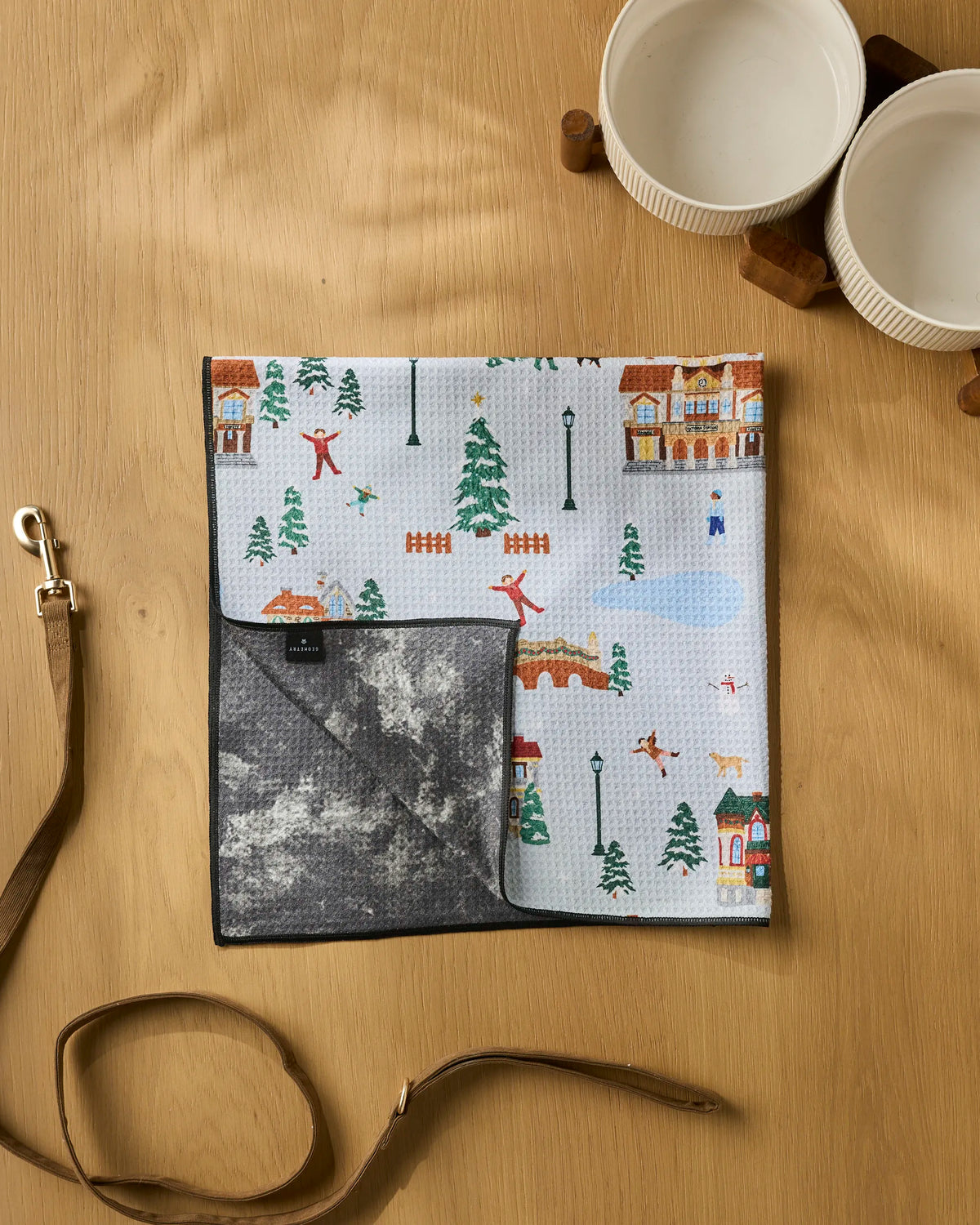 Holiday Nostalgia by Geometry—a highly absorbent dog towel made from recycled materials featuring a holiday-themed winter village print—paired with a dog leash and two white pet bowls on a wooden surface.