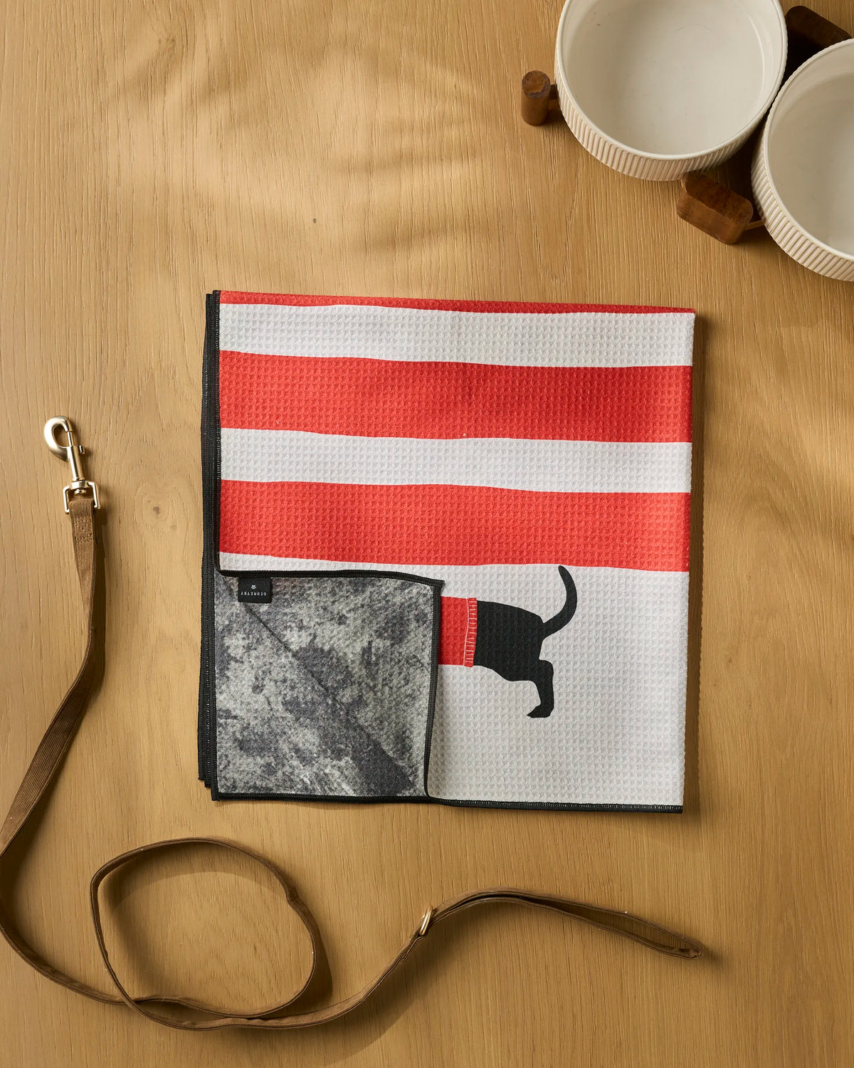 A dog leash, two white bowls on a wooden stand, and the Geometry Holiday Stretch dog towel—featuring a black dog illustration and crafted from highly absorbent, recycled materials—are displayed on a wooden surface.