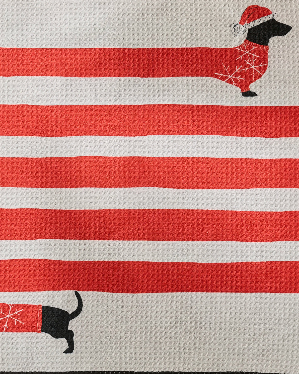 Introducing the “Holiday Stretch” towel by Geometry—a festive, eco-friendly towel crafted from recycled materials. Featuring bold red and white stripes, it is adorned with playful black dachshund illustrations dressed in cozy Santa hats and sweaters, with just their heads and front legs peeking over the top edge and tails with back legs along the bottom. Highly absorbent and designed to add a touch of holiday cheer to your space, this unique towel combines joyful style with sustainability.
