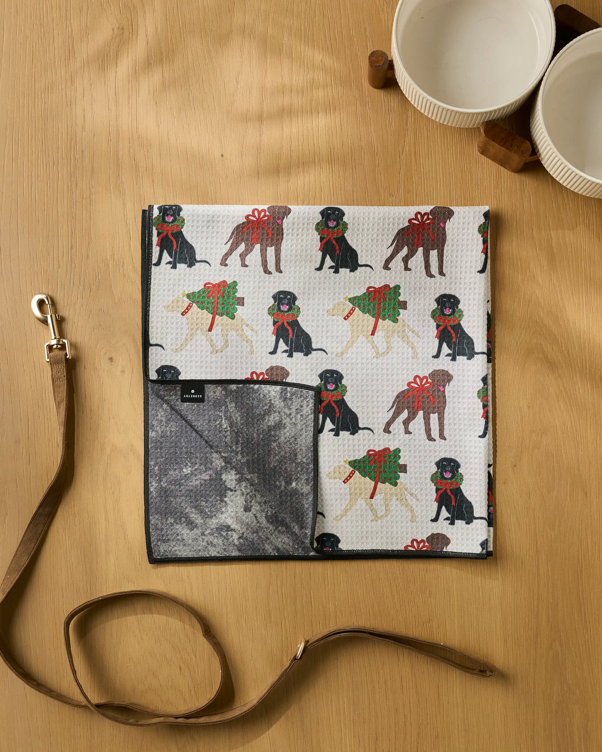 A folded, highly absorbent Geometry Merry Labs dog towel featuring illustrated dogs with Christmas wreaths, alongside a brown leather leash and two empty white bowls on a light wood surface.