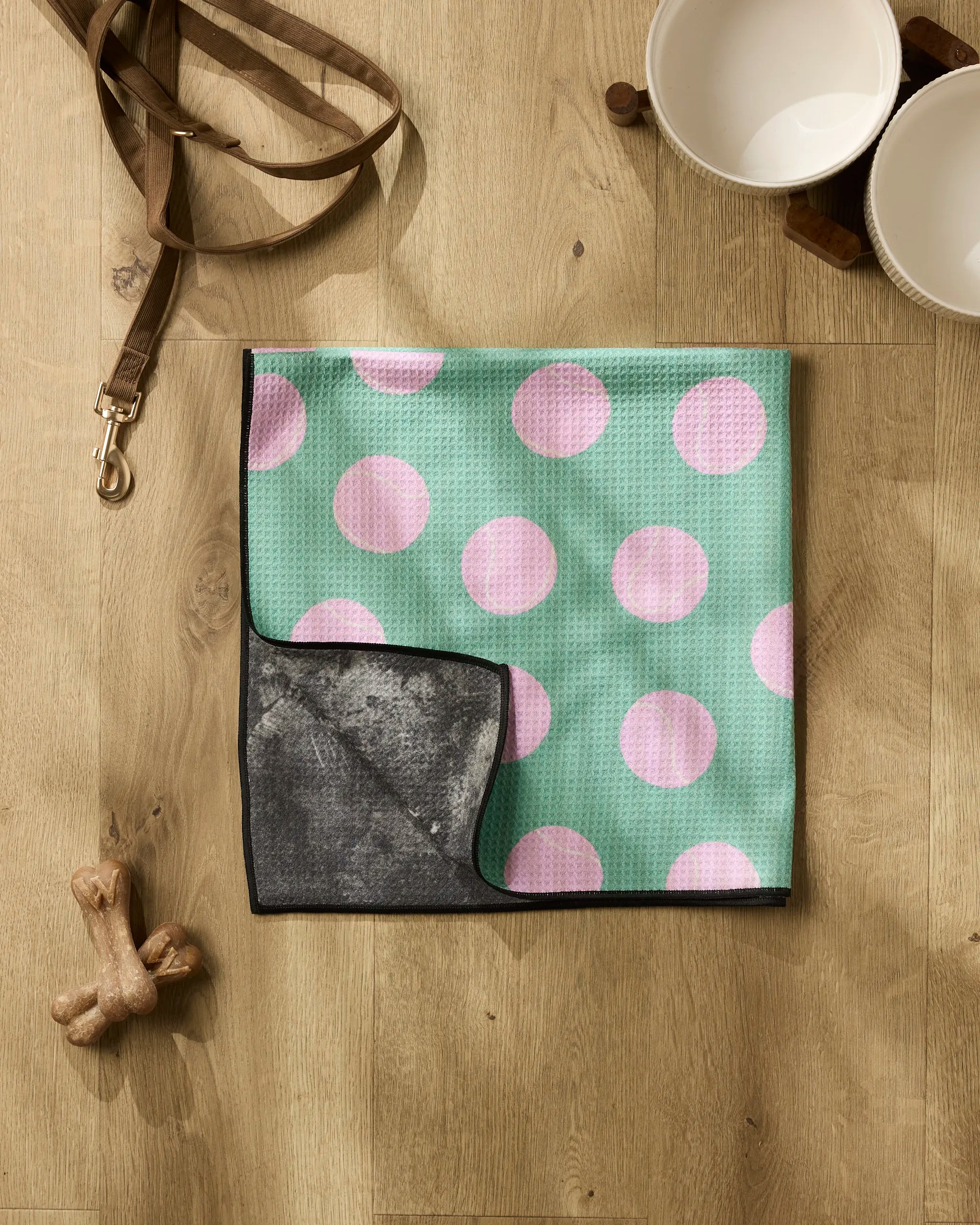 A Geometry Pink Ball rests on a wooden floor beside two white pet bowls on a stand, a brown leash, and two dog bone chew toys.