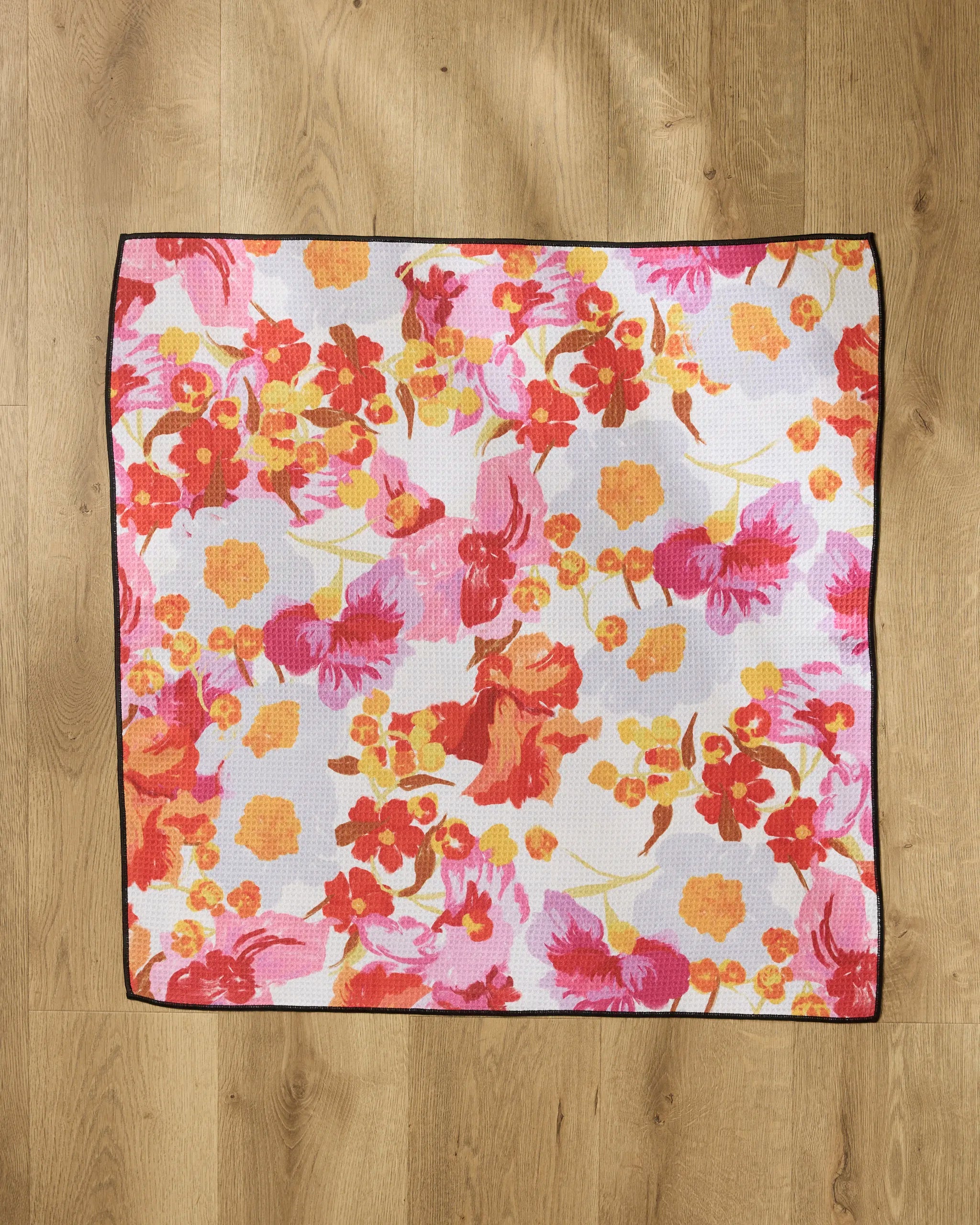 The Geometry Pup Florals dog towel, made from recycled materials, showcases a vibrant orange, red, and pink floral design on white with a thin black border and is displayed flat on a light wooden floor.