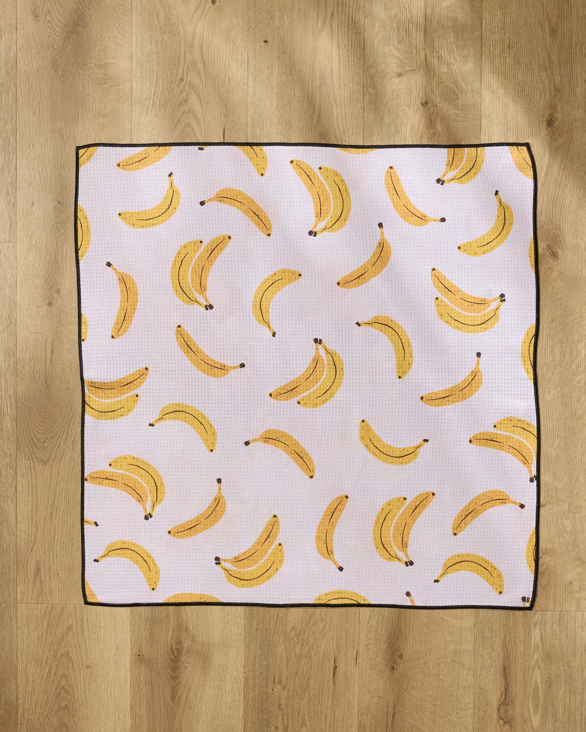 The Geometry Pupnana is a square, highly absorbent dog towel made from recycled materials, featuring a playful yellow banana pattern on white with a black border, shown flat on a light wooden floor.