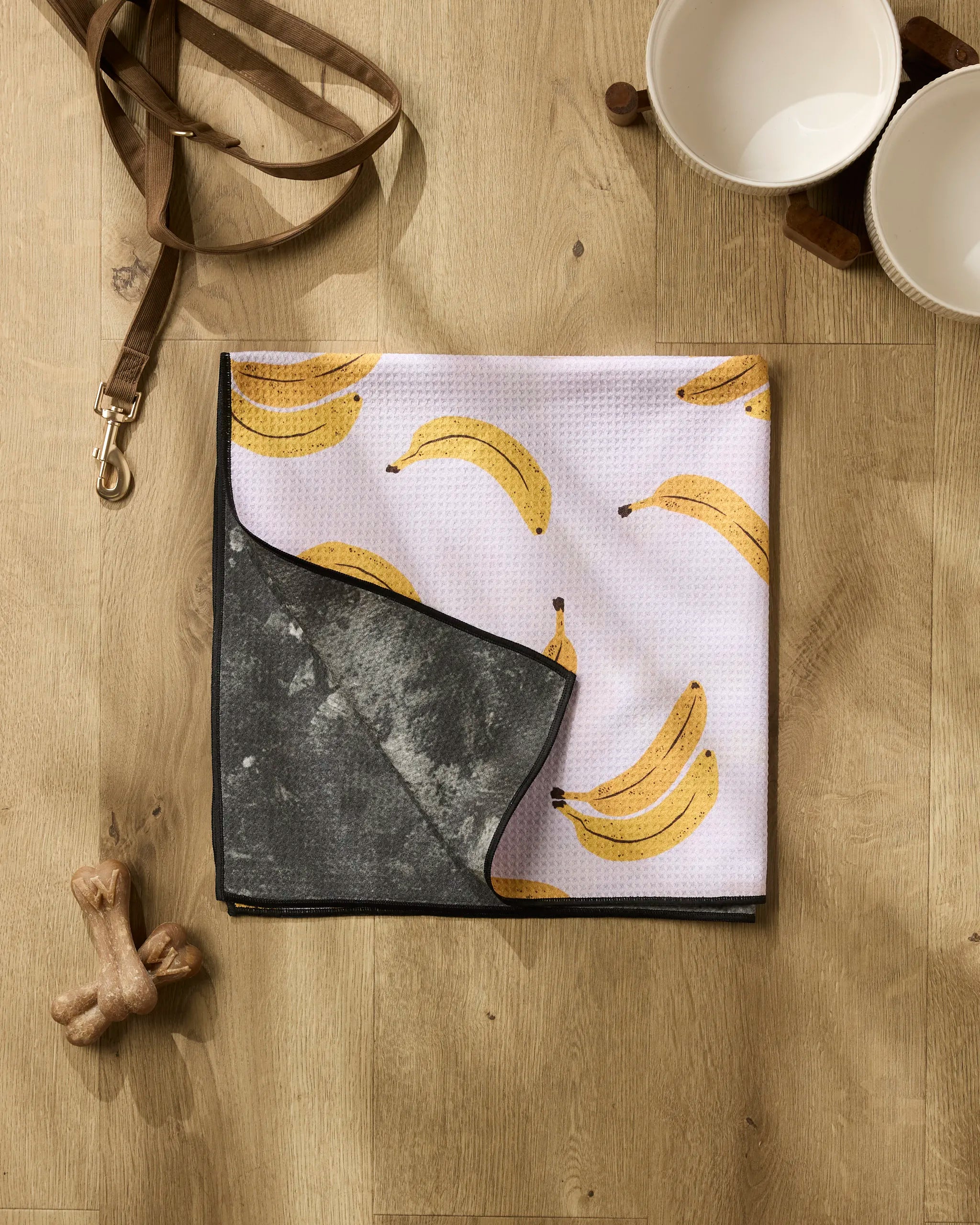 A dog leash, two white bowls, and two dog bones sit on a wooden floor next to the Geometry Pupnana dog towel, crafted from recycled materials with a banana print and dark underside for high absorbency.
