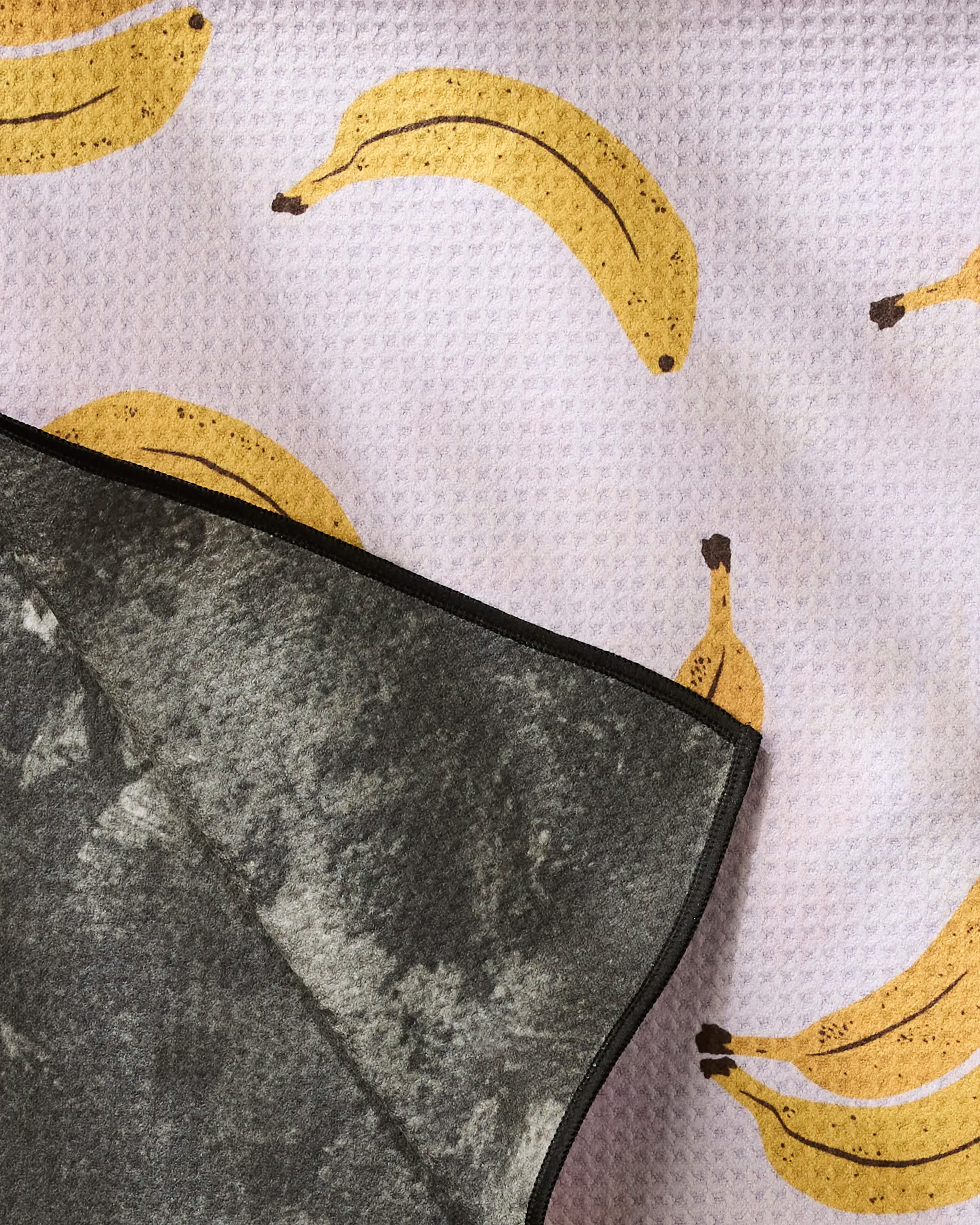 The Geometry Pupnana dog towel, featuring a marble-like design and high absorbency, partially covers a textured fabric patterned with yellow bananas on a light background.