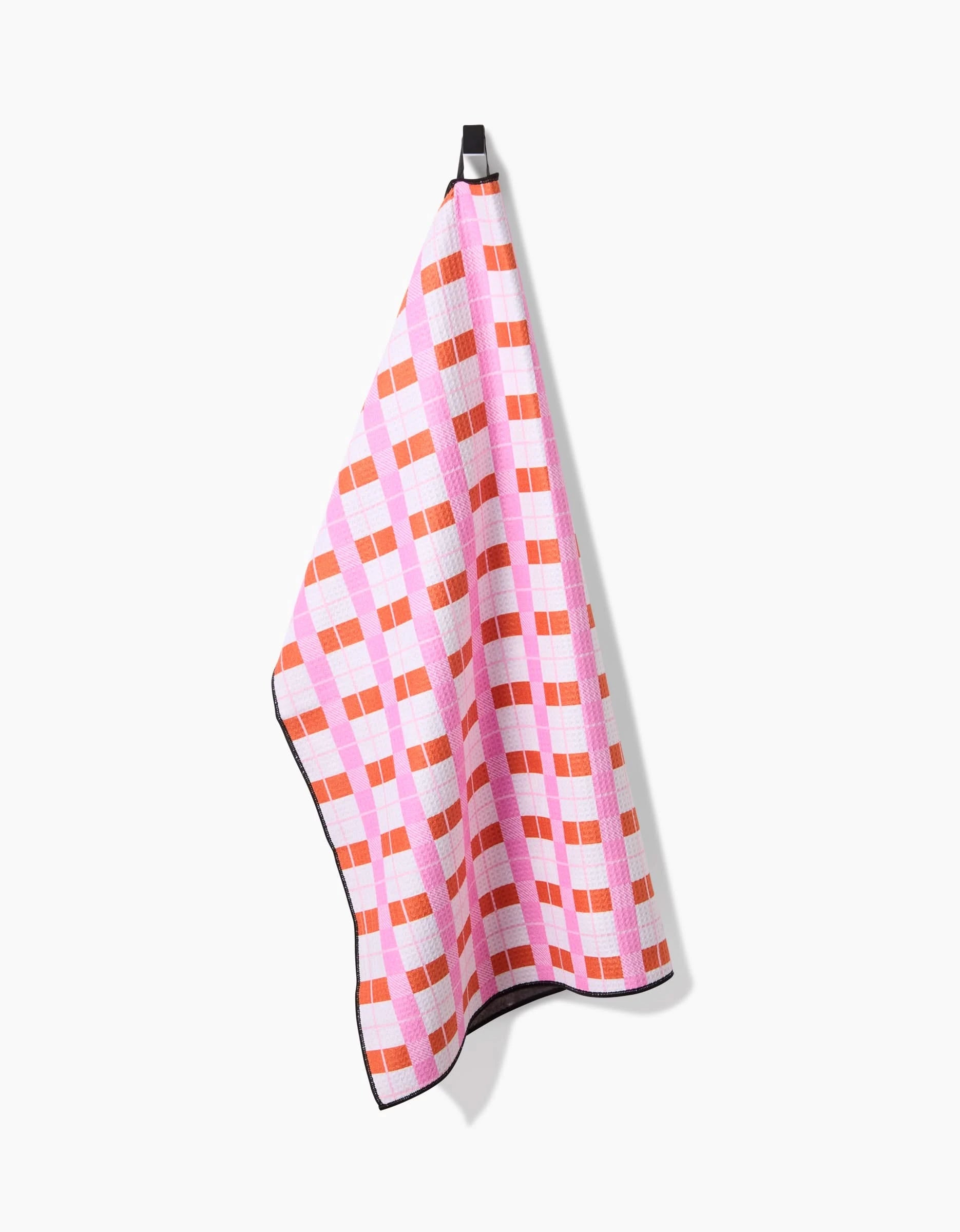 The Megan Roy Red Pink Plaid towel, featuring a pink, red, and white checkered design, hangs from a black loop against a plain white background.