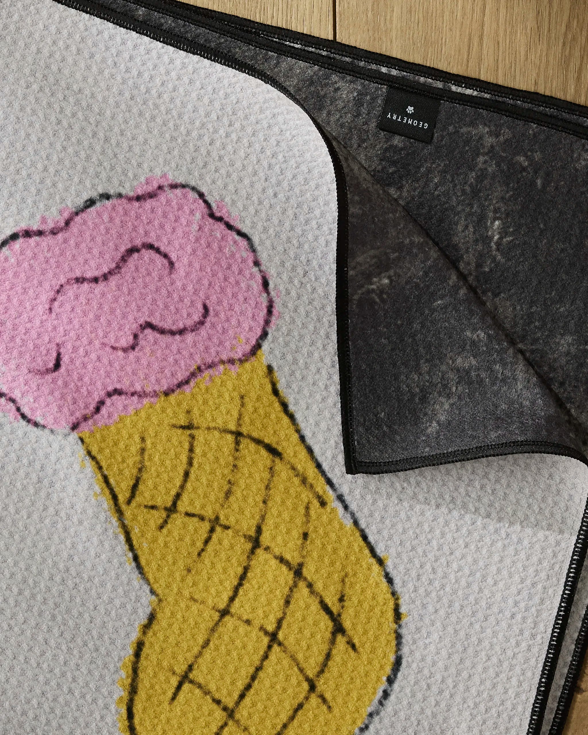 A close-up of Geometry's Silly Stockings doormat shows a pink ice cream scoop in a yellow cone on recycled material, with one corner flipped to reveal the black, non-slip backing. The mat rests on a wooden floor.