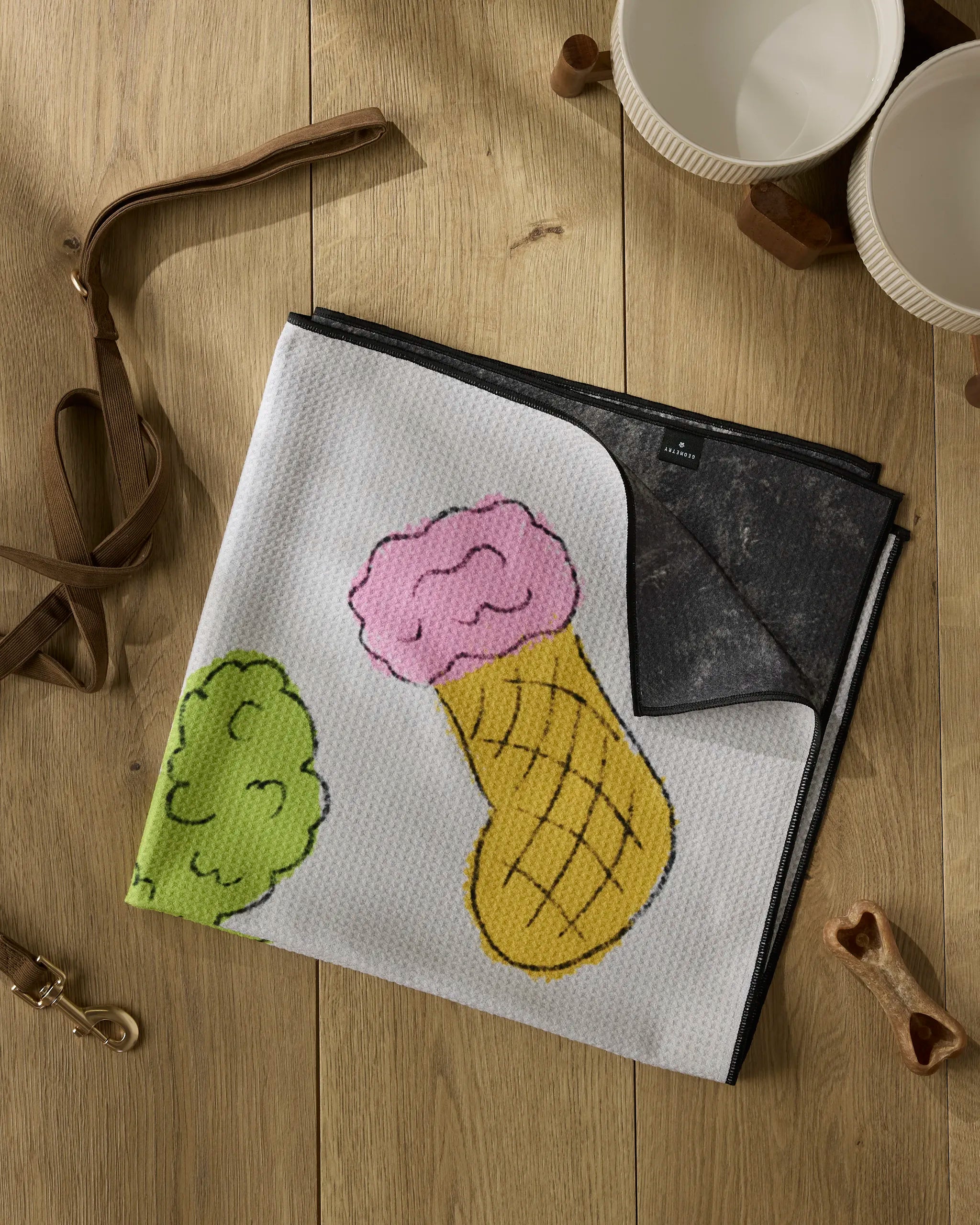The Silly Stockings dog towel by Geometry, made from recycled materials and featuring colorful ice cream cone and green scoop illustrations, is folded on a wooden floor beside two white bowls, a brown leash, and a dog bone toy.