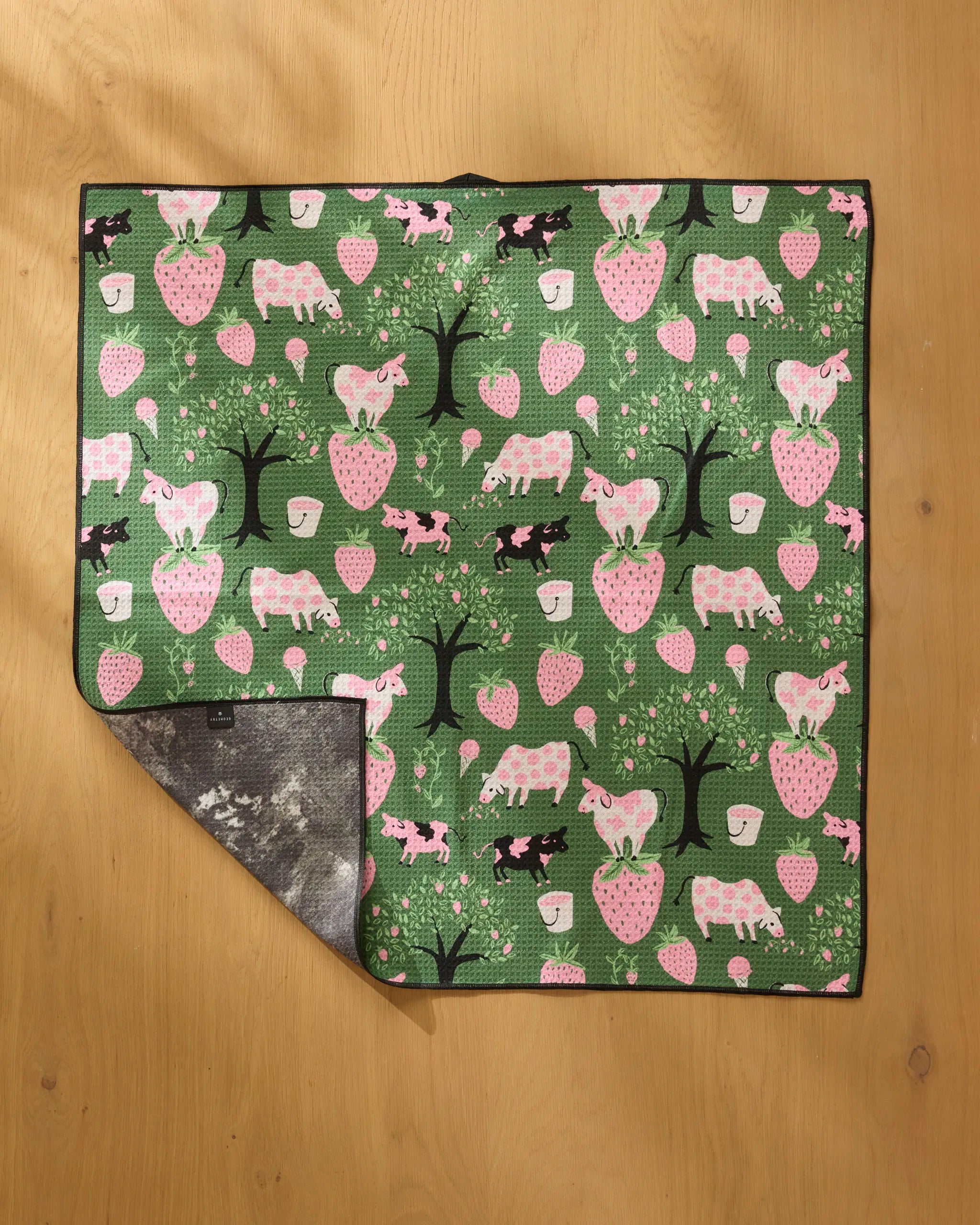 The “Strawberry Cows” mat by Louise Pretzel features pink and white strawberries, trees, unicorns, cows, and hedgehogs in a green repeating pattern. Made from recycled materials with a gray underside visible on the folded corner.